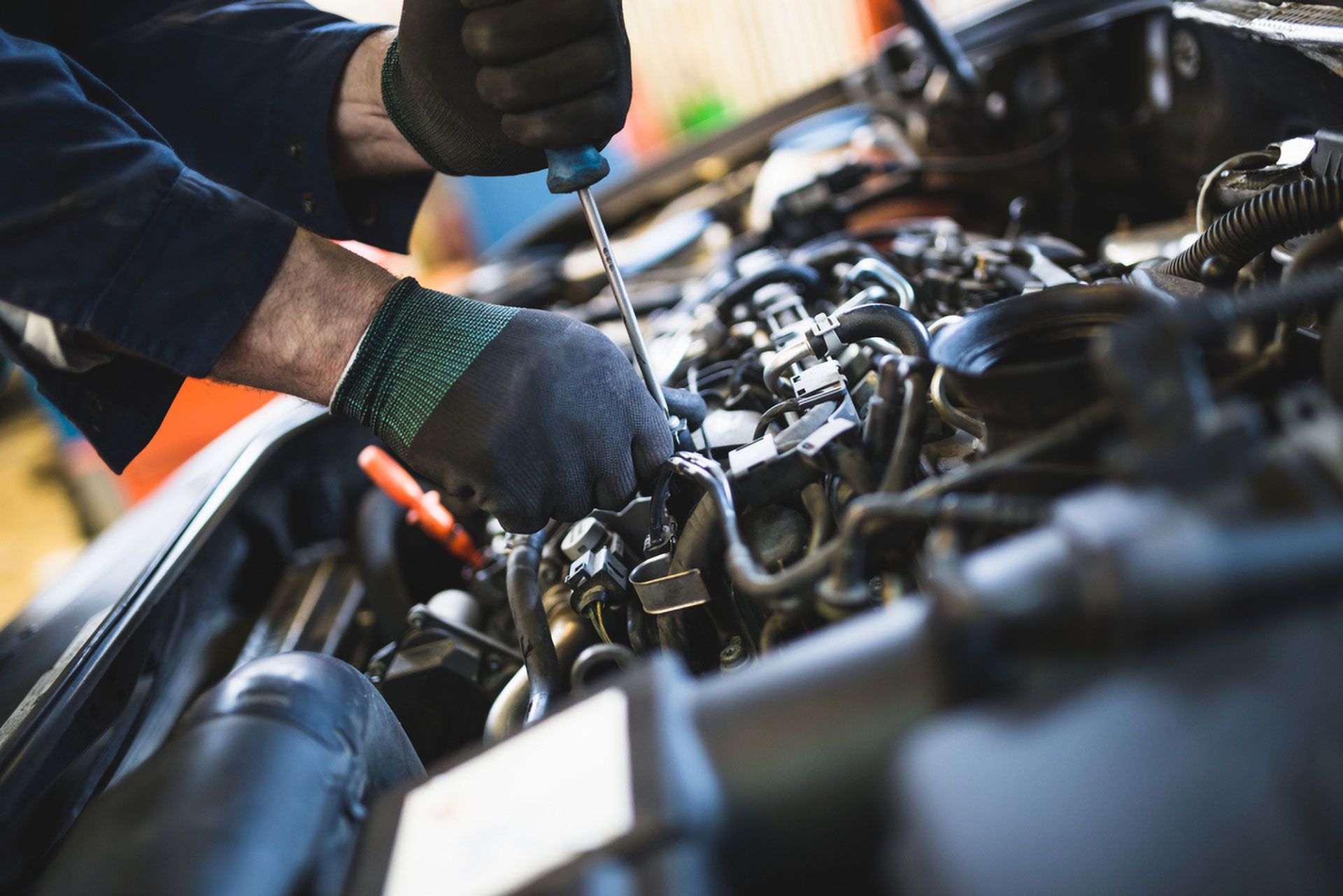 Mechanic wearing dark gloves uses a long screwdriver to work on a car engine.