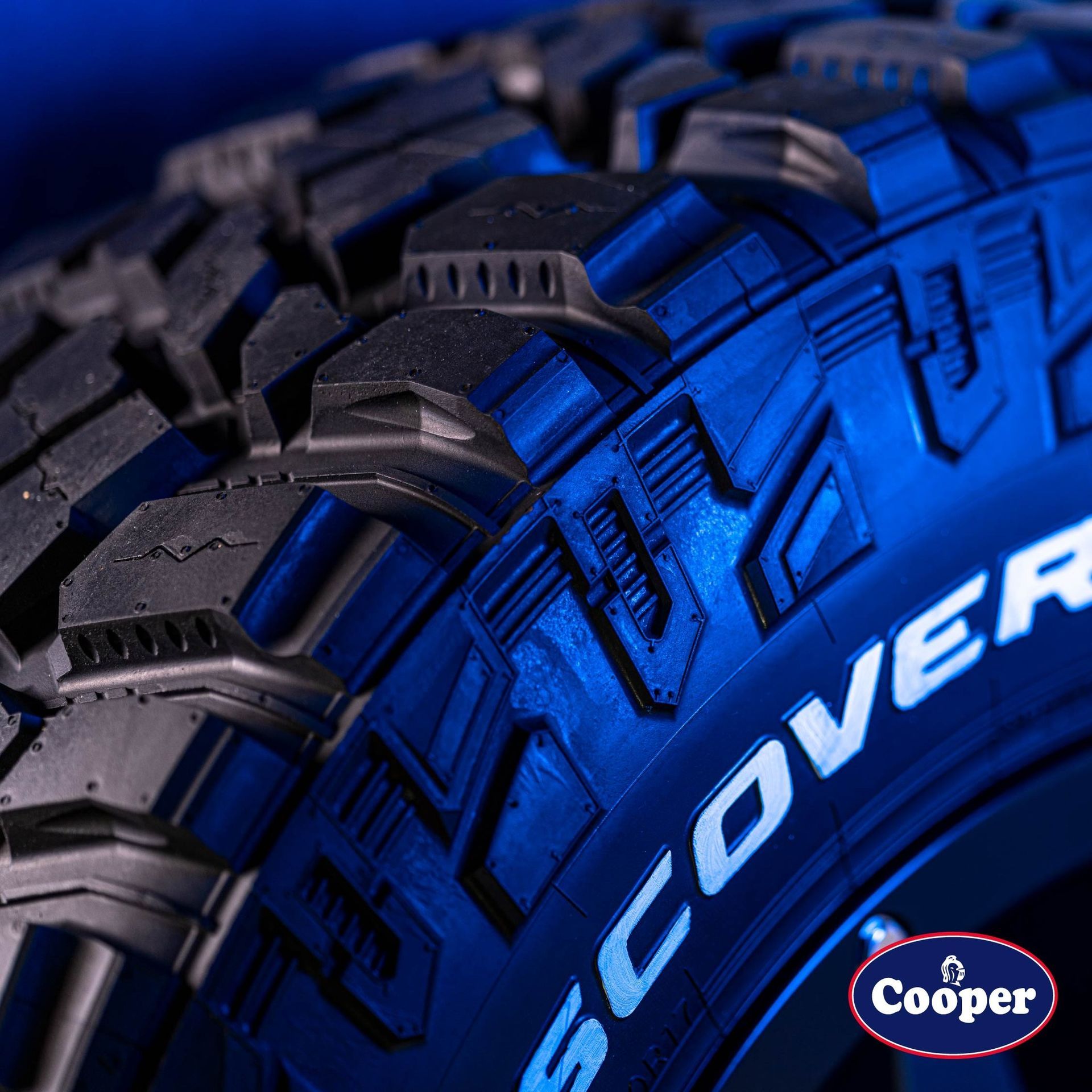 Close-up of a Cooper Discoverer tire with an aggressive tread pattern, illuminated in blue light against a dark background.