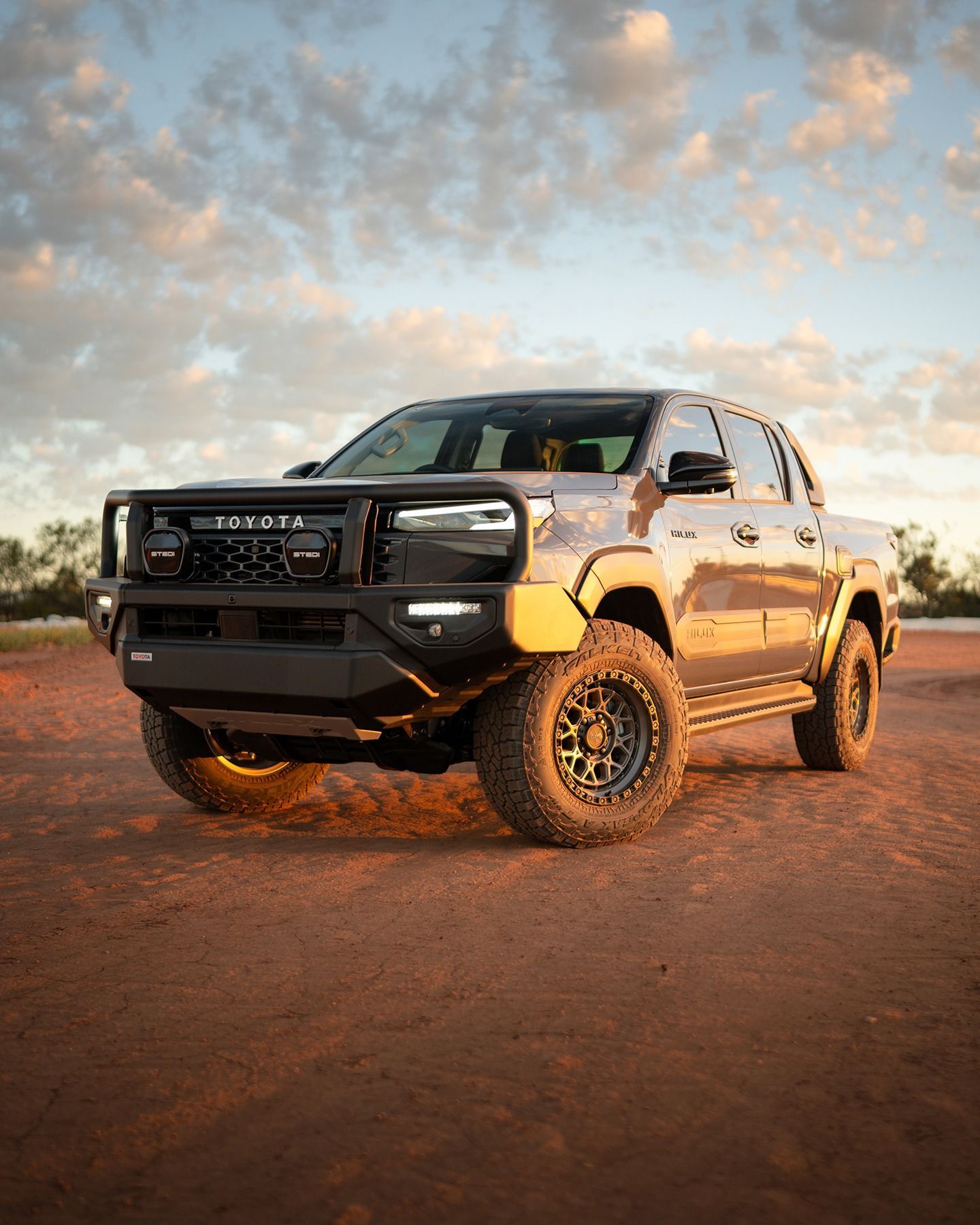 A grey Toyota pickup truck with a black off-road bull bar and large tires parked on reddish dirt at sunset.