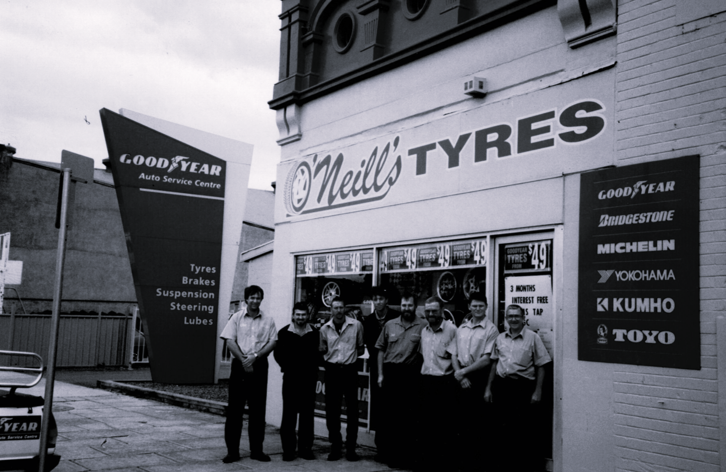 A black-and-white photo of seven workers standing in front of O’Neill’s Tyres shop with a Goodyear sign in the background.