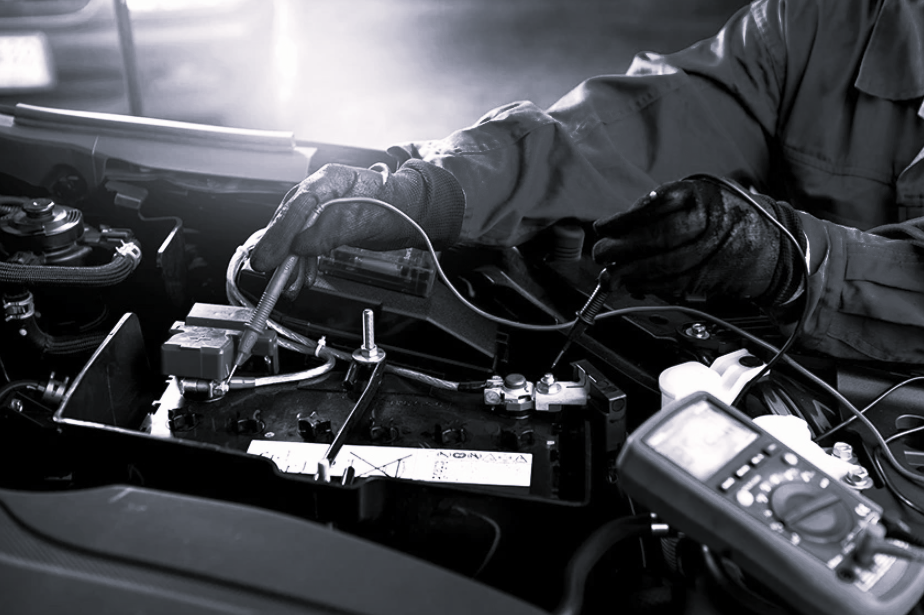 A mechanic in gloves uses a multimeter to test a car battery under the hood of a vehicle.