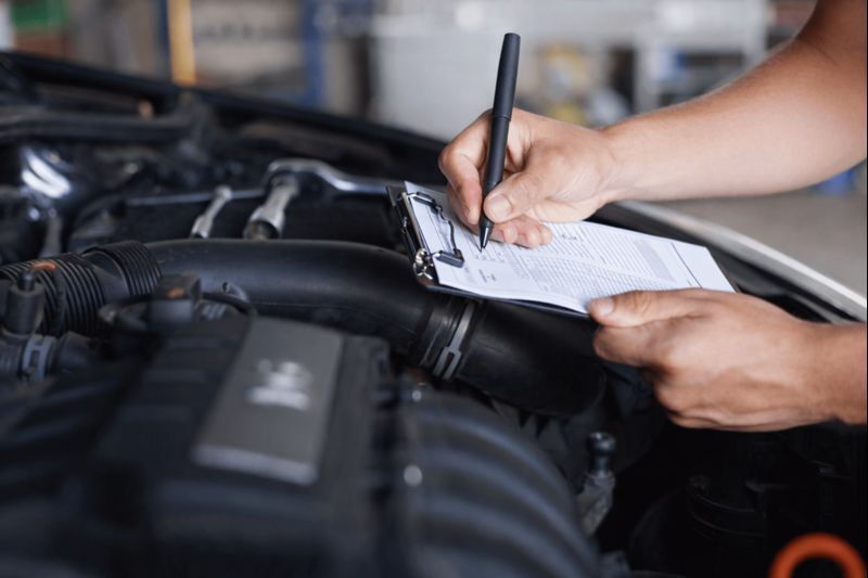 A mechanic writes on a clipboard while performing an inspection under the hood of a car.
