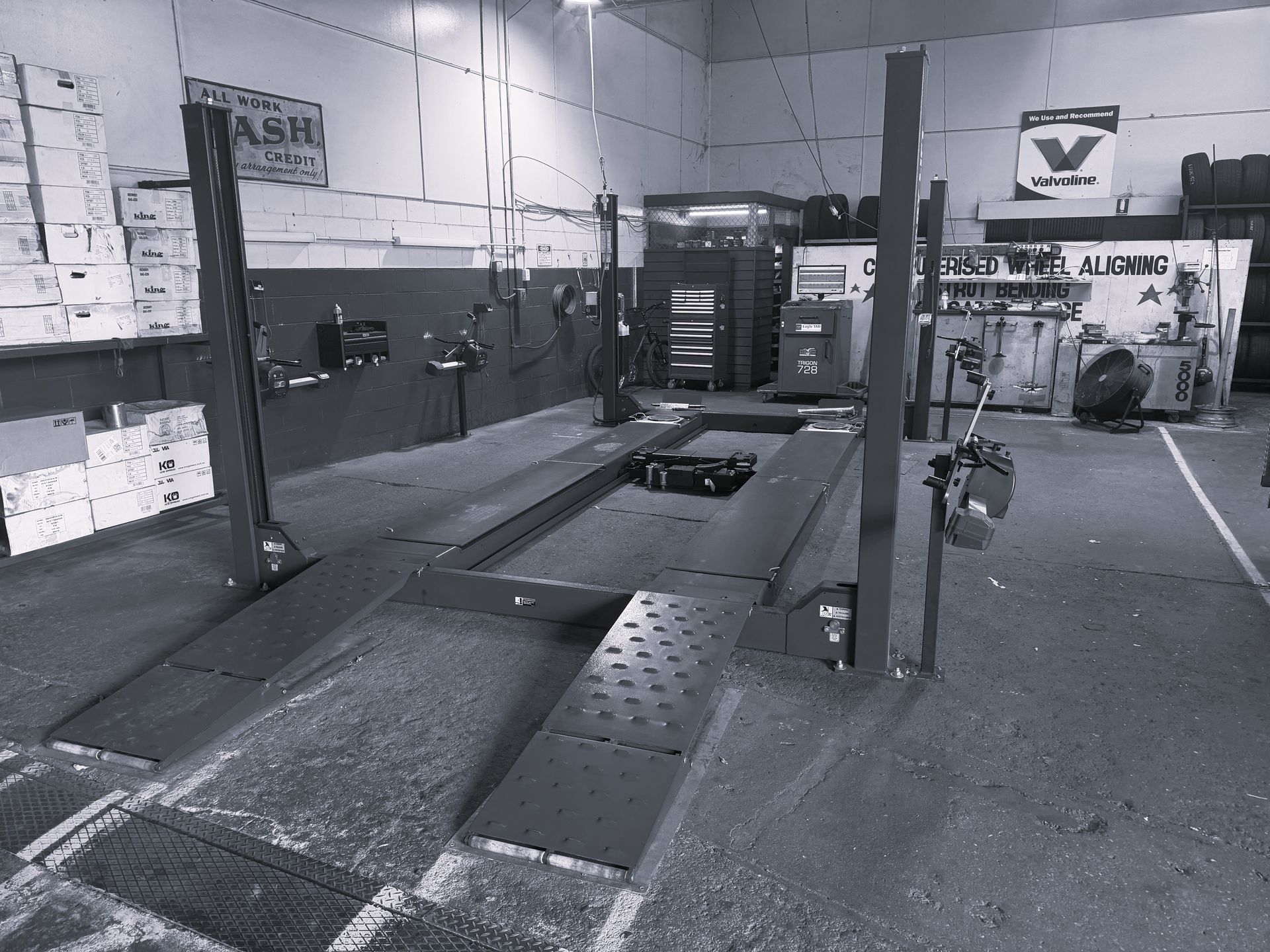 An empty automotive repair lift stands in a workshop with stacks of boxes, tools, and tires in the background.