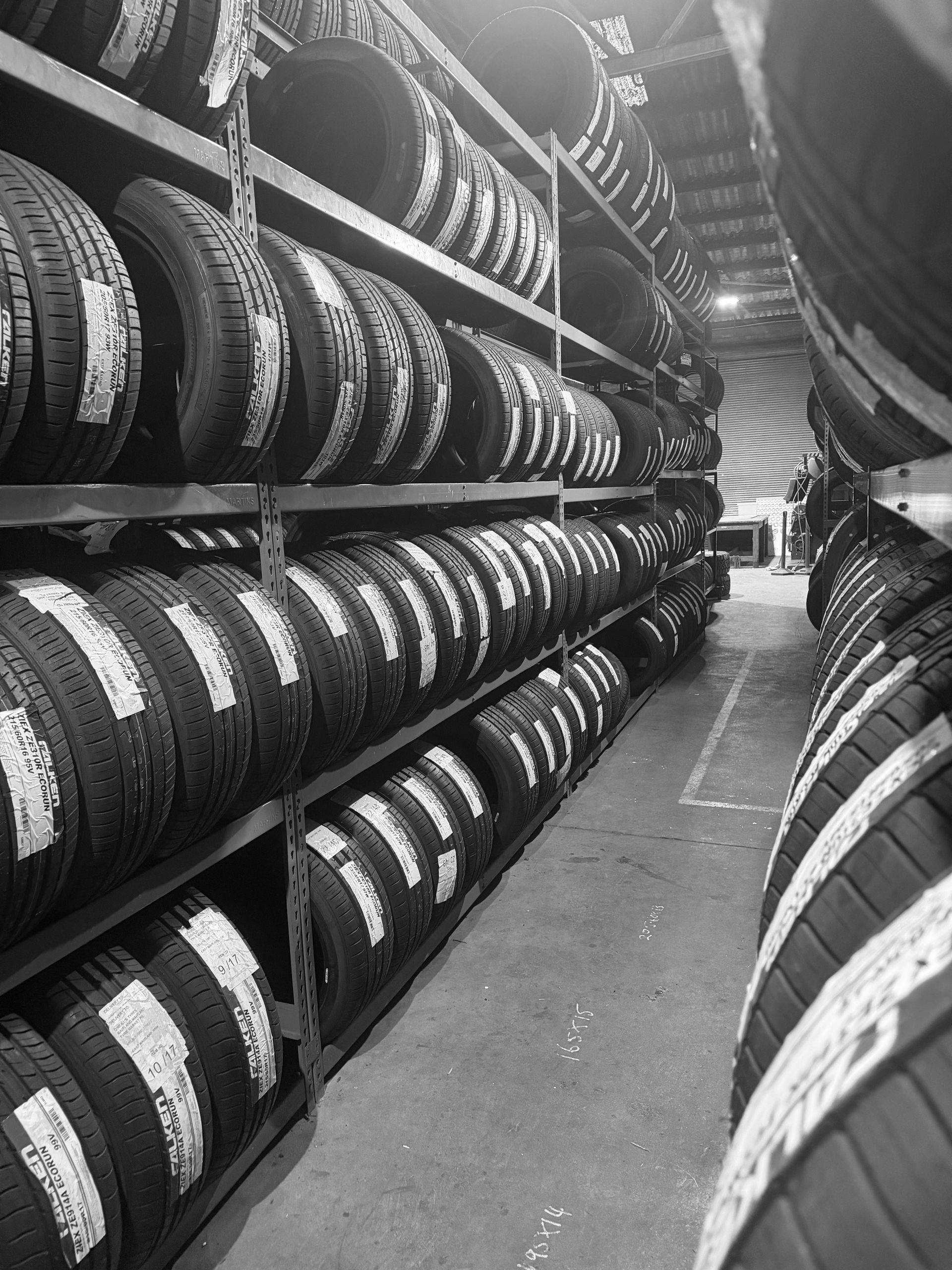 Black and white photo of a warehouse aisle filled with rows of tires stacked on metal shelving units.