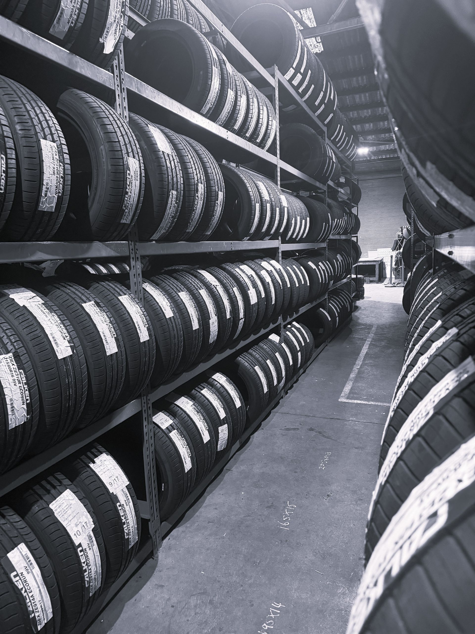 Black and white image of a warehouse aisle with rows of car tires neatly stacked on tall metal industrial shelving.