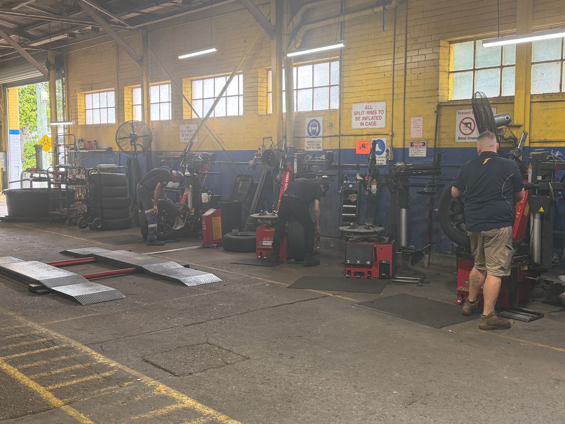Two mechanics work on tires in a spacious, yellow-walled industrial workshop with large windows.