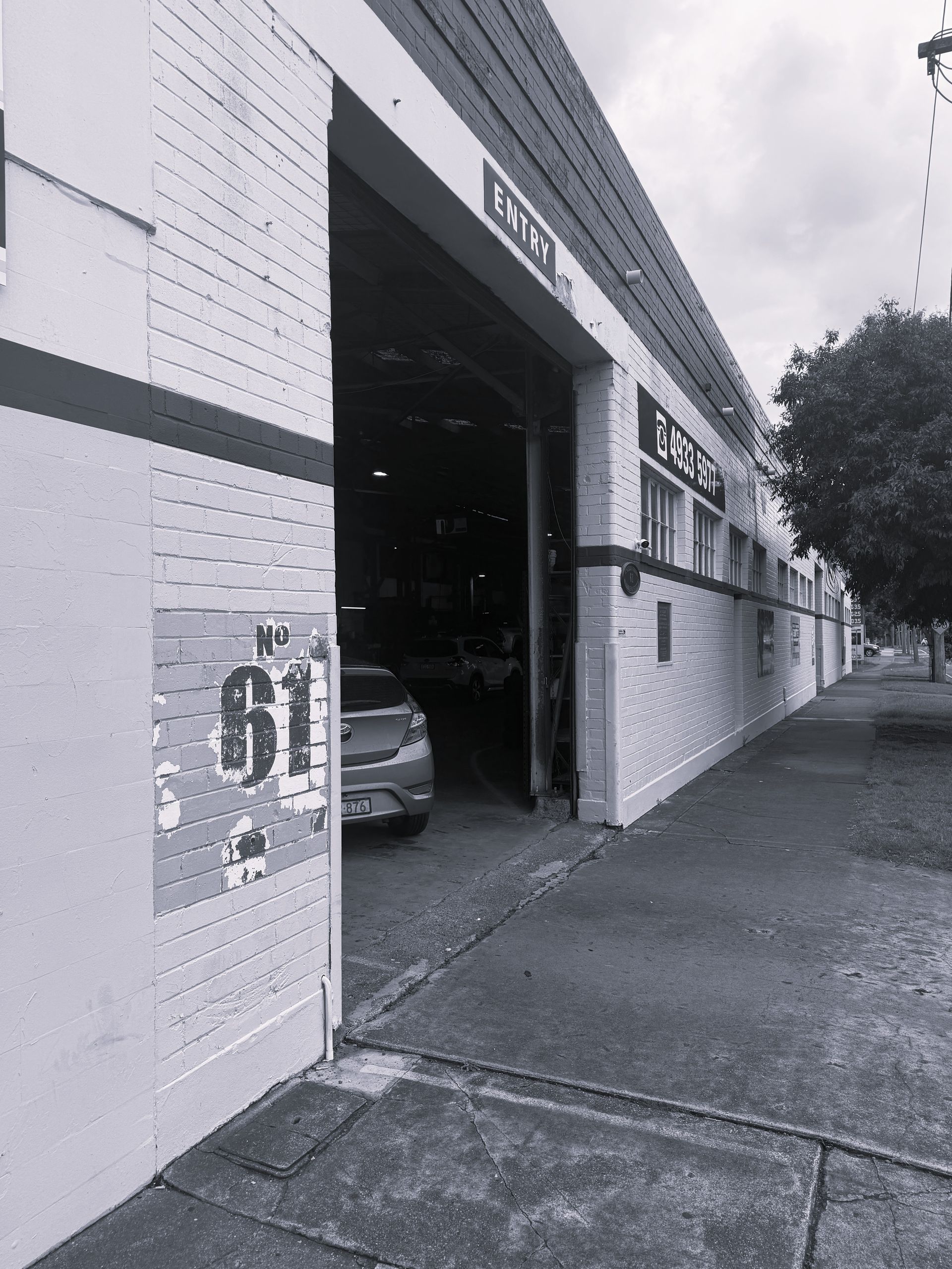 A sidewalk passes a white brick building with a large open garage bay revealing a parked car and the number 81 on the wall.