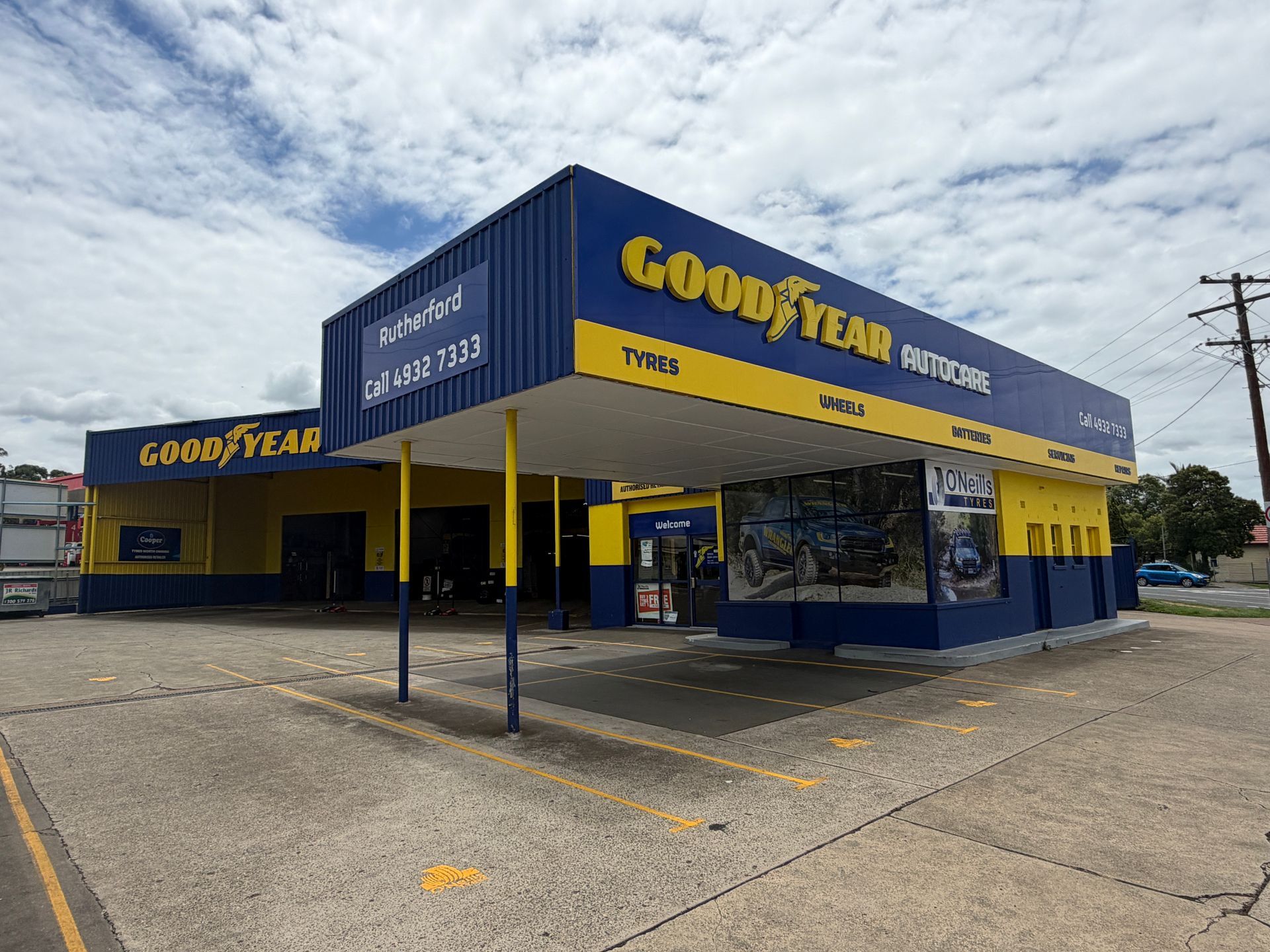 O'Neills Tyres storefront with a yellow facade, blue trim, and a black signage pillar on a paved lot under a cloudy sky.