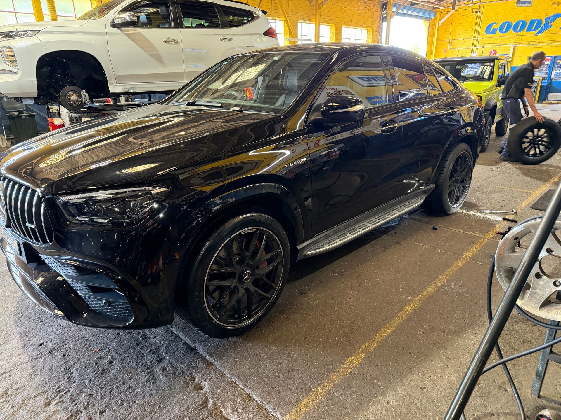 A black luxury SUV parked in an auto repair shop with a mechanic working on a tire in the background.