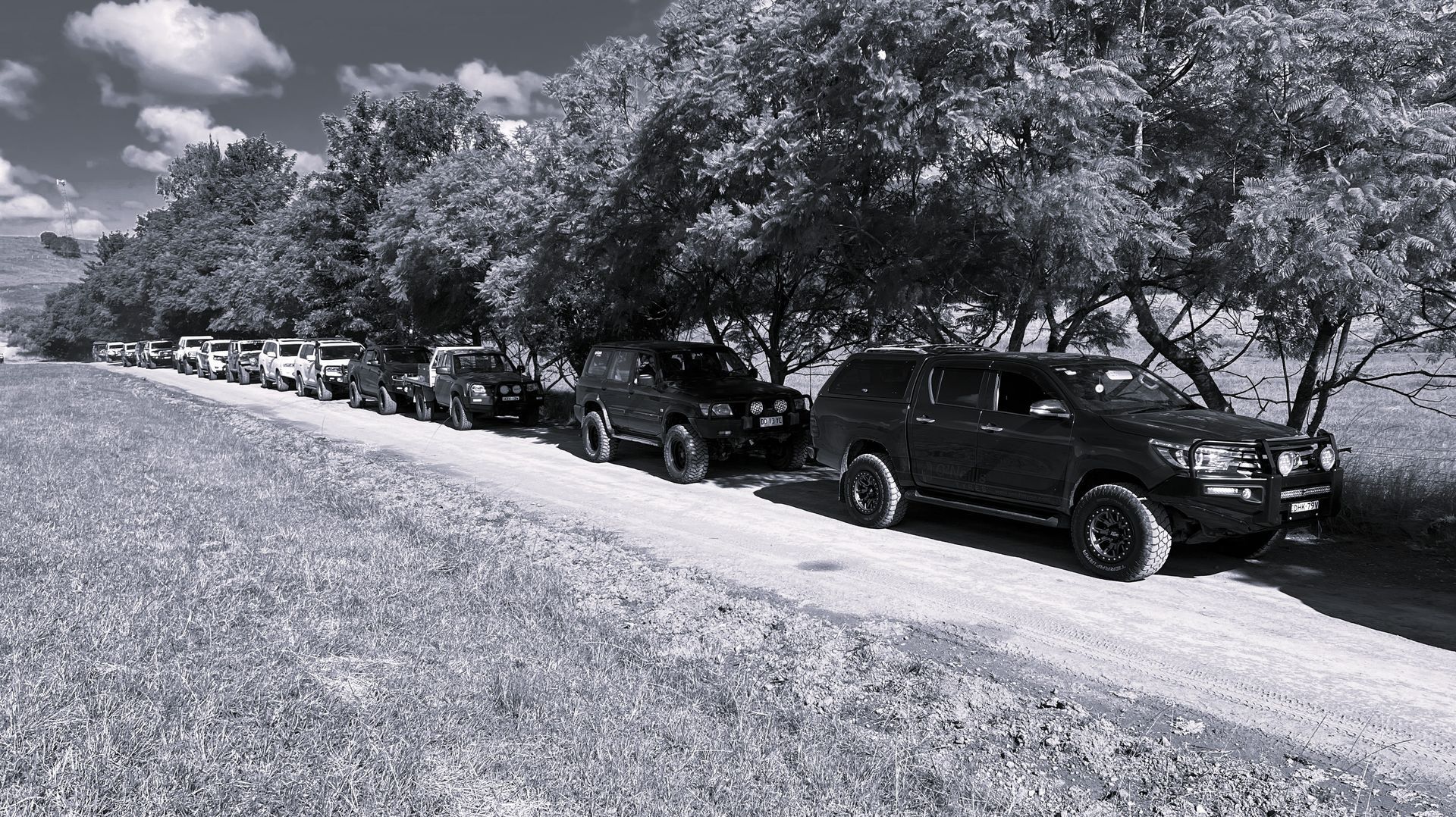 A line of dark off-road vehicles parked on a dirt road beside a row of trees under a cloudy sky.