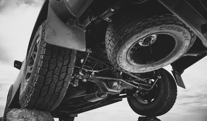 A low-angle, grayscale view of an off-road vehicle's undercarriage, showing tires resting on rocks and exposed suspension.