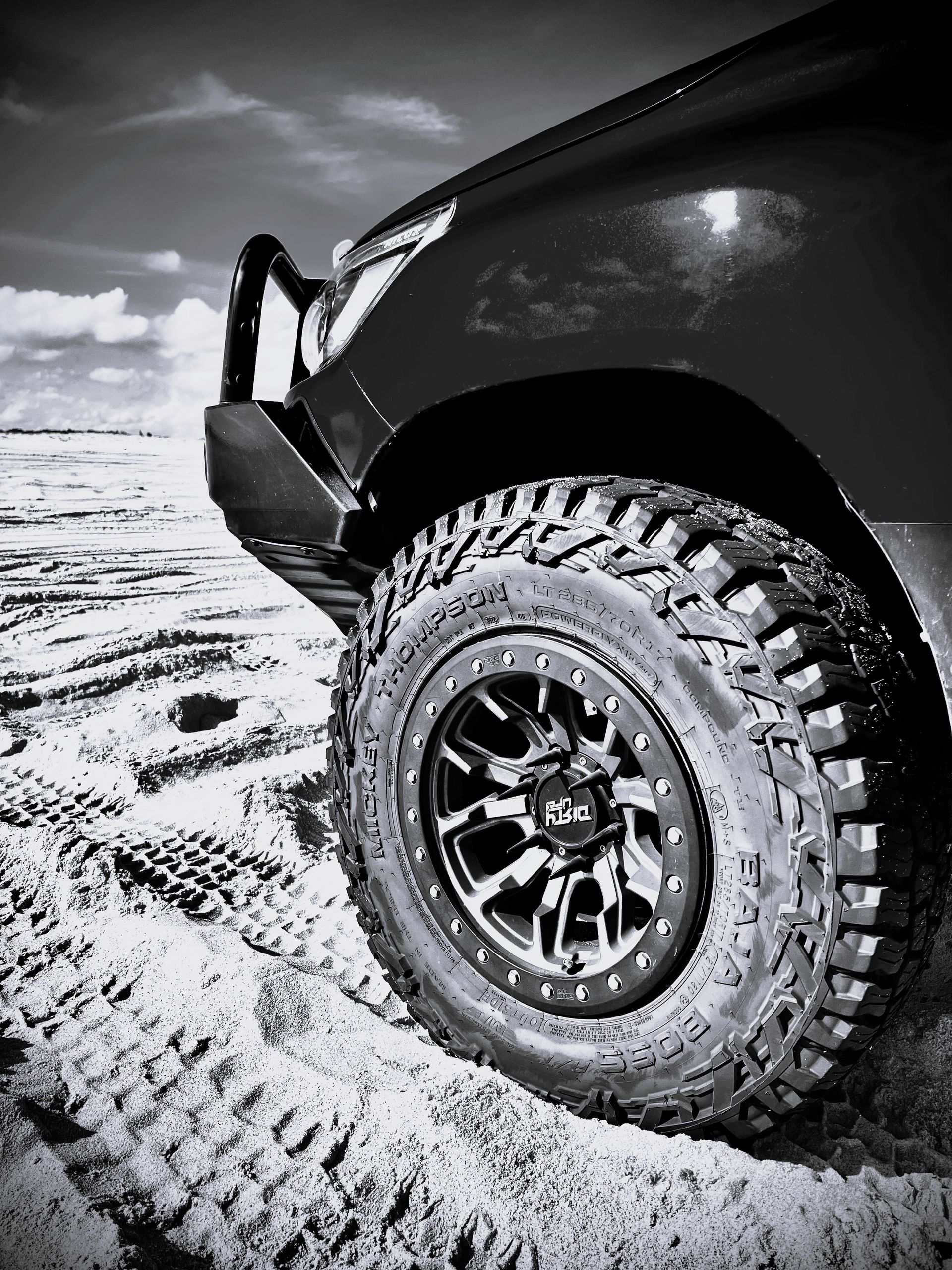 A black-and-white low-angle close-up of a rugged off-road vehicle tire and heavy-duty bumper on a sandy track.