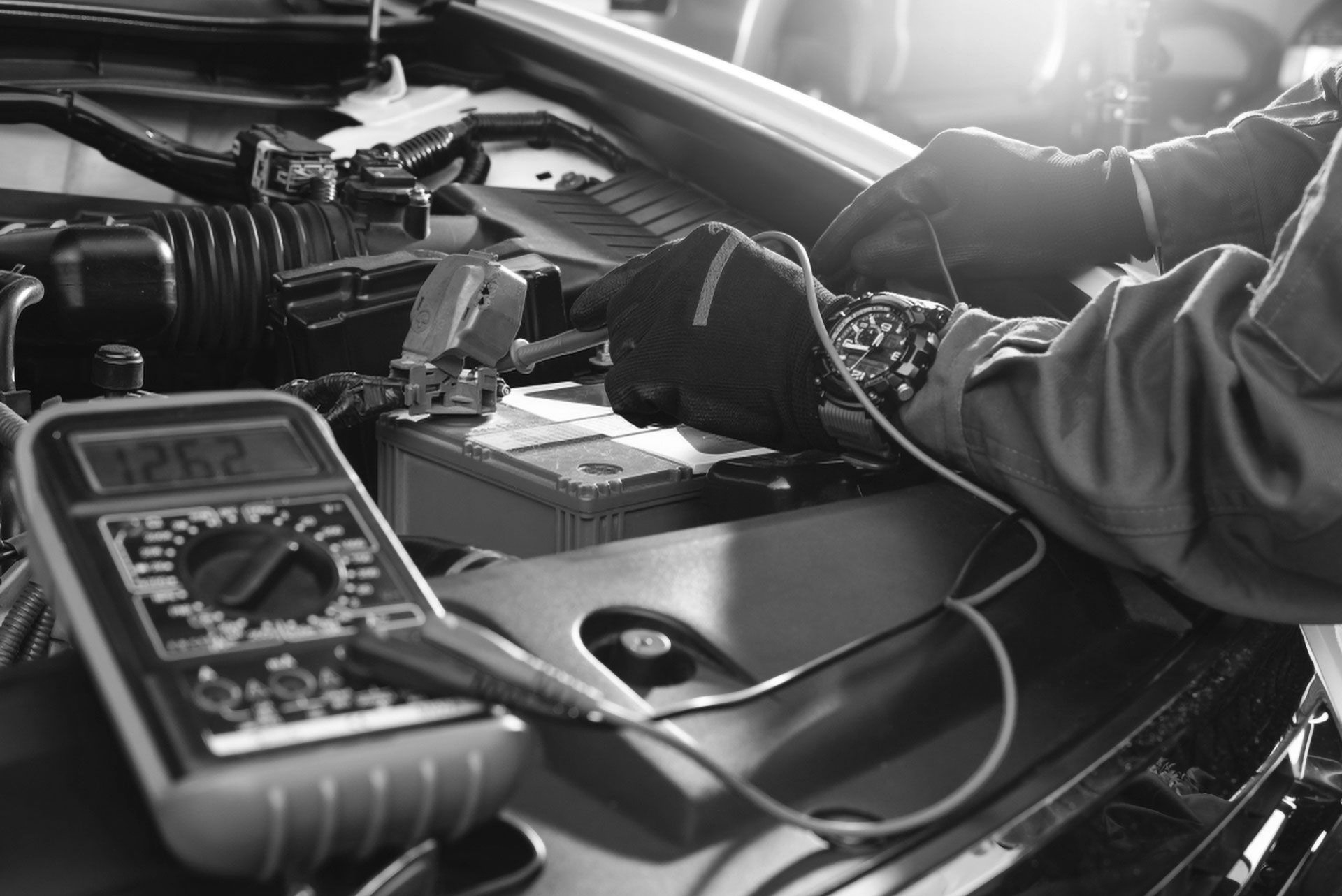 A mechanic wears black gloves while using a multimeter to test a car battery under the hood.