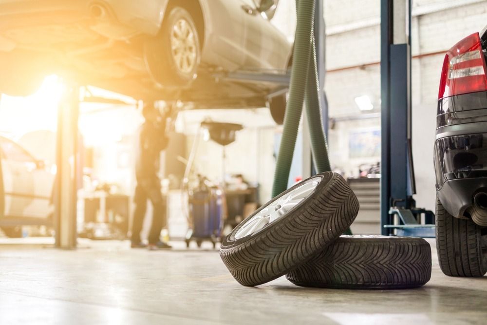 A mechanic works under a car lifted on a hoist in a garage, with spare tires resting on the floor in the foreground.