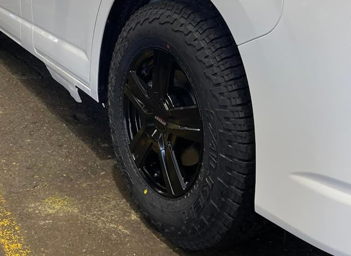 A close-up view of a black alloy wheel with an all-terrain tire mounted on a white vehicle.