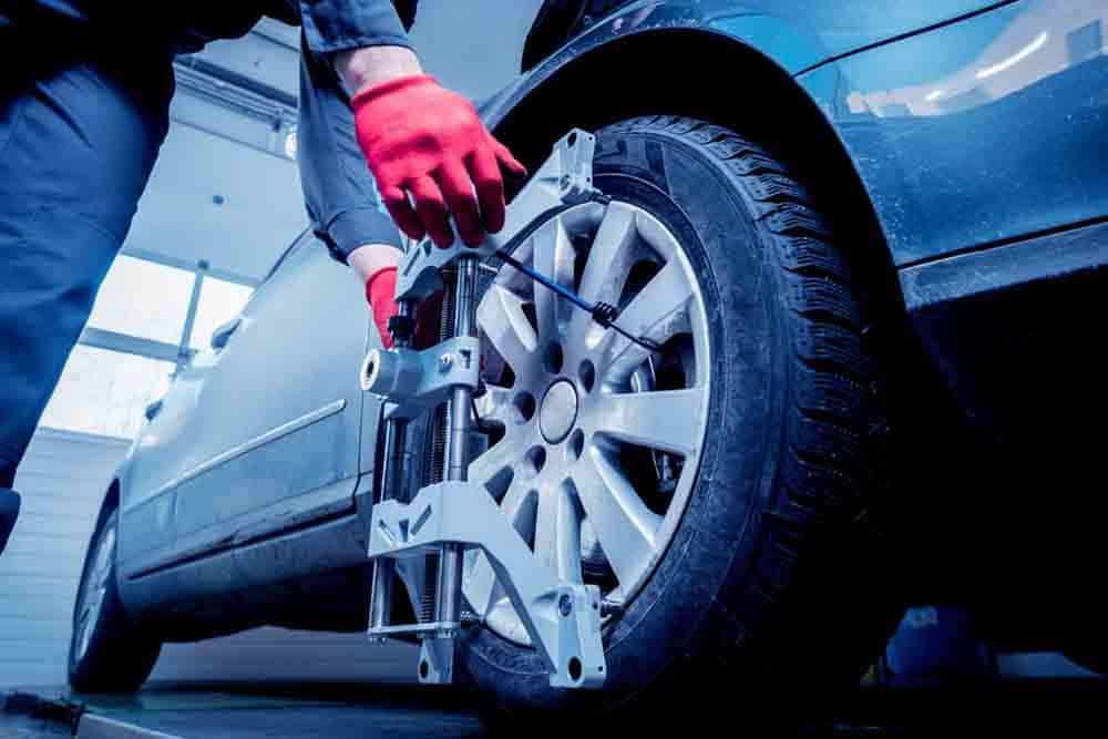 A mechanic in red gloves attaches a wheel alignment sensor to the front tire of a car in a repair shop.