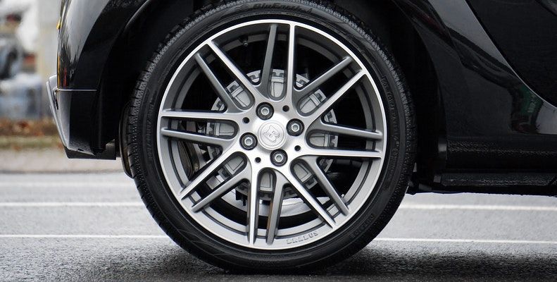 A close-up of a black car's multi-spoke silver alloy wheel and tire on a paved surface.