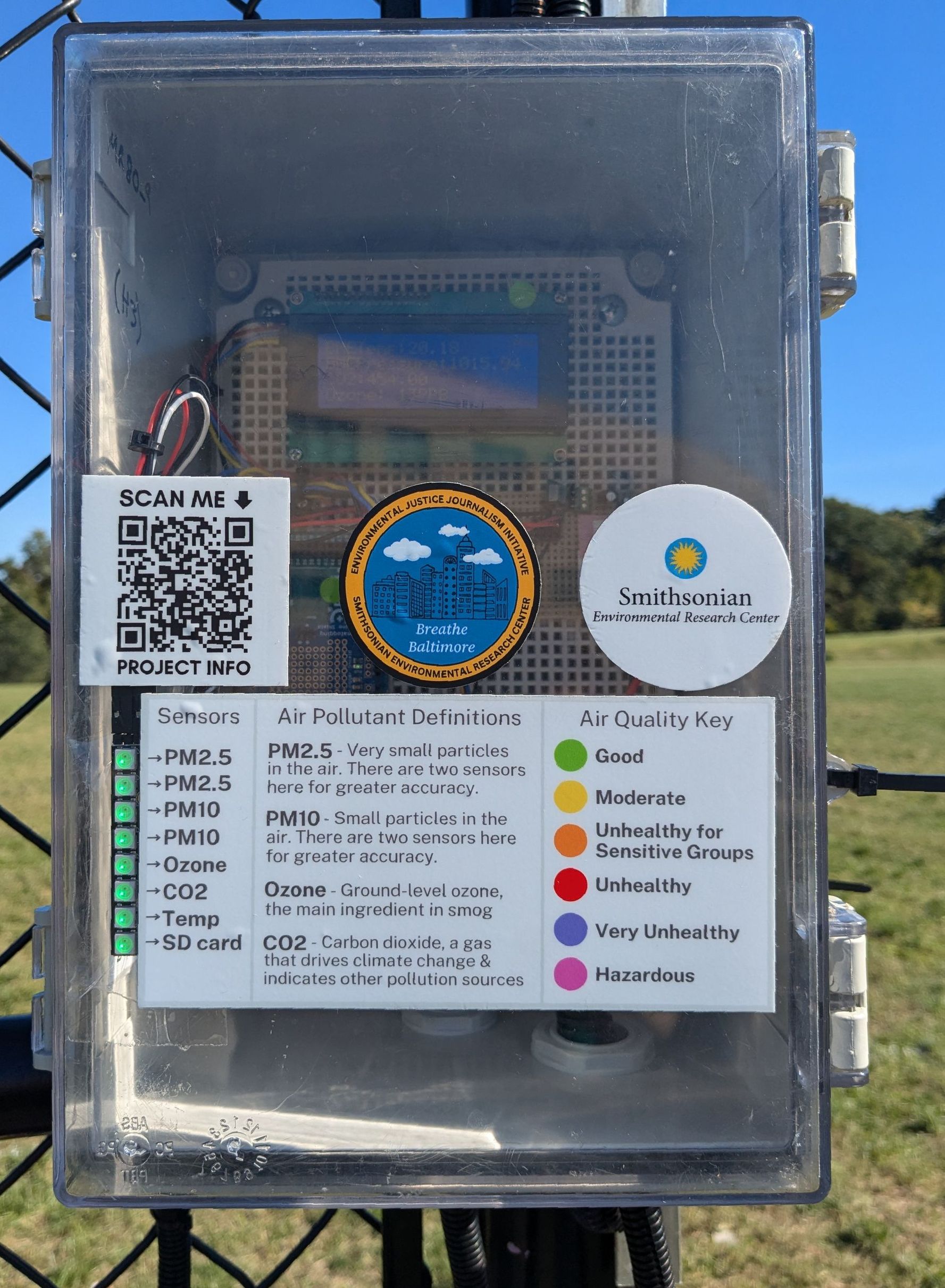 Image of a gray box with a clear door cover mounted onto the end of a black chain-link fence. The box houses a green sensor inside with a blue LED screen and different color wires. On the door are 4 stickers; one with a QR code, a Breathe Baltimore logo, a SERC logo, and a sticker with air pollution definitions. To the left of the stickers is an LED light strip that changes color based the air quality.