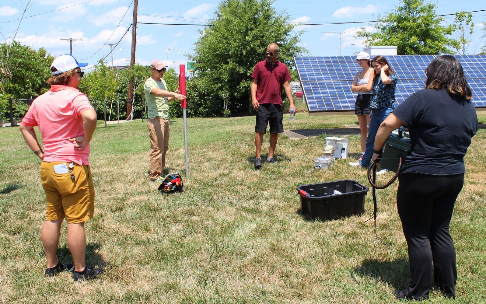 5 people gather around a spot in the grass to identify where they are going to put pilot air quality sensor. It is a sunny day with blue skies and some clouds. Behind them is a larger solar panel and trees. The people are holding different tools.