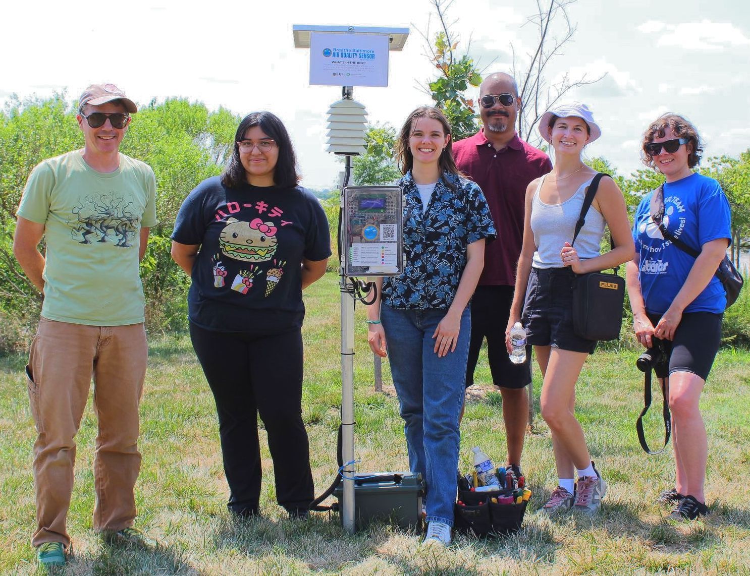 6 people, men and women, stand on both sides of the air quality sensor. The sensor is mounted on a pole with a solar panel on top.