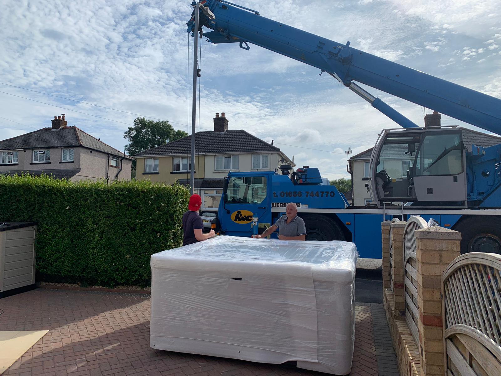 A blue crane is lifting a hot tub in a driveway.