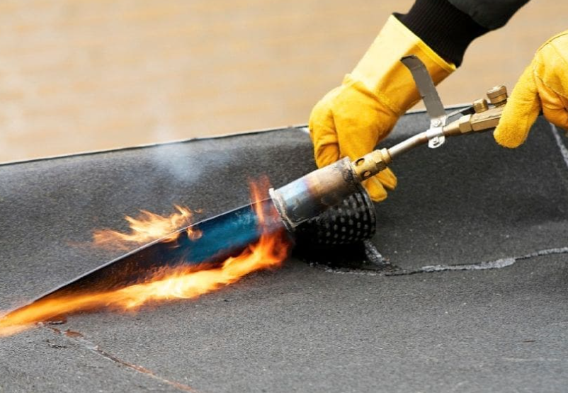 Un operaio addetto alla copertura utilizza una fiamma per sigillare il materiale del tetto. I guanti gialli proteggono le mani dalla fiamma.