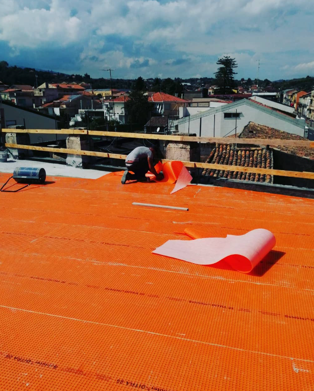 Persona che stende del materiale di copertura arancione su un tetto, con uno sfondo di paesaggio urbano.