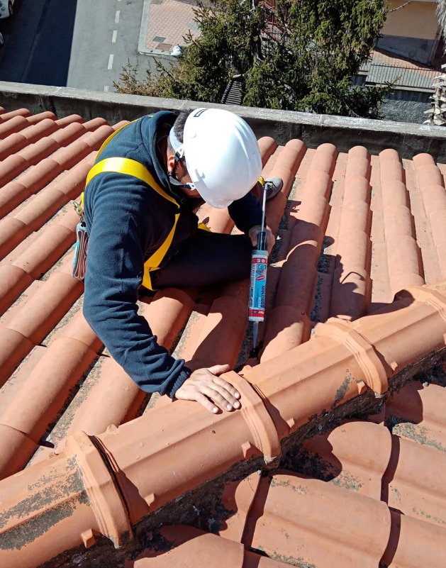 Un operaio edile con indosso l'equipaggiamento di sicurezza sigilla le tegole con una pistola per calafataggio, in una giornata di sole.
