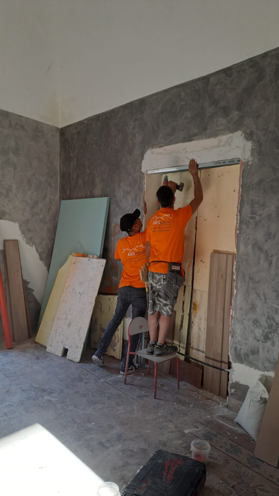 Due operai con le camicie arancioni installano pannelli di legno attorno a una porta in una stanza con pareti grezze.