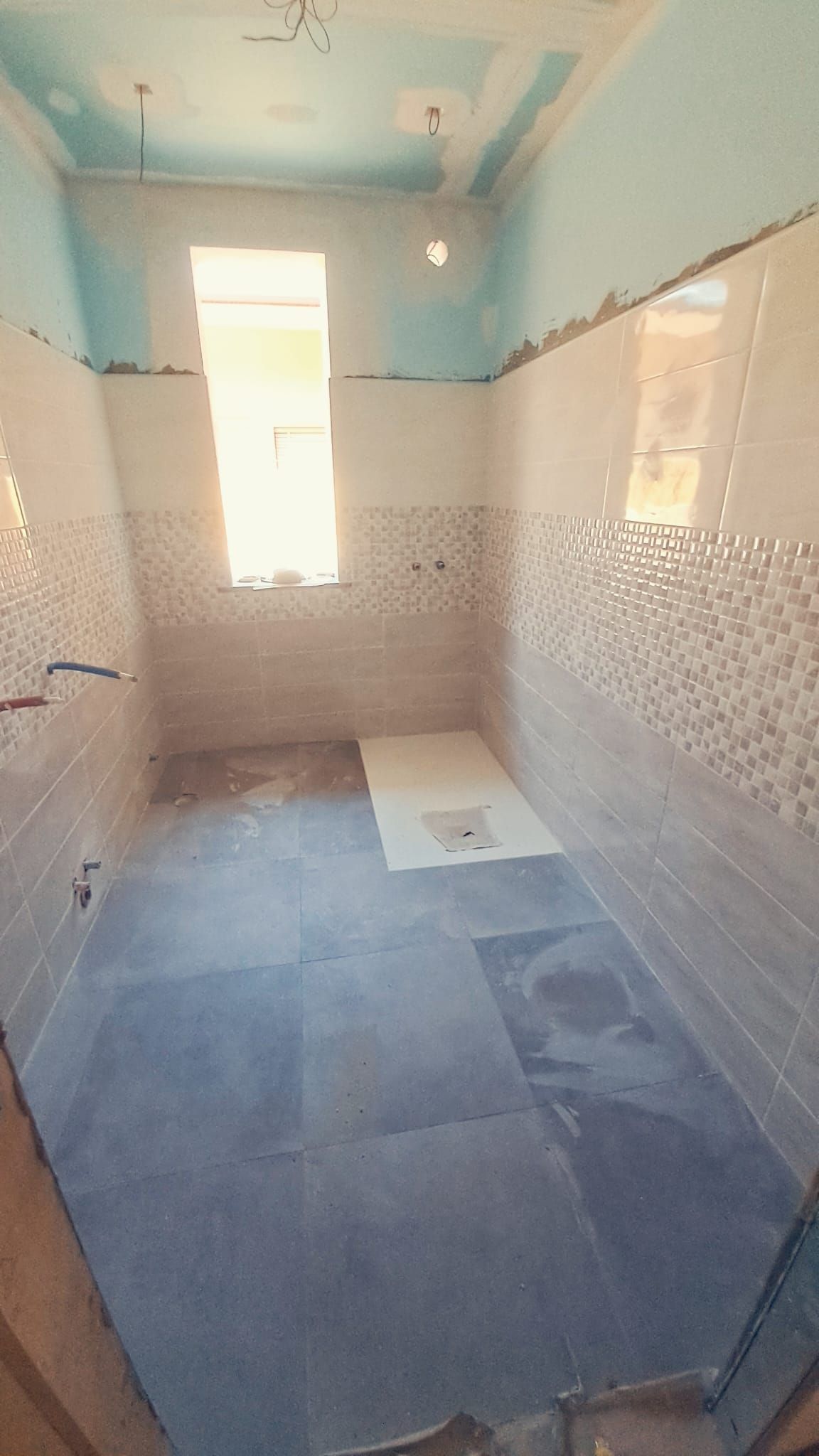 Bagno in costruzione, con pareti piastrellate, soffitto grezzo e una finestra.