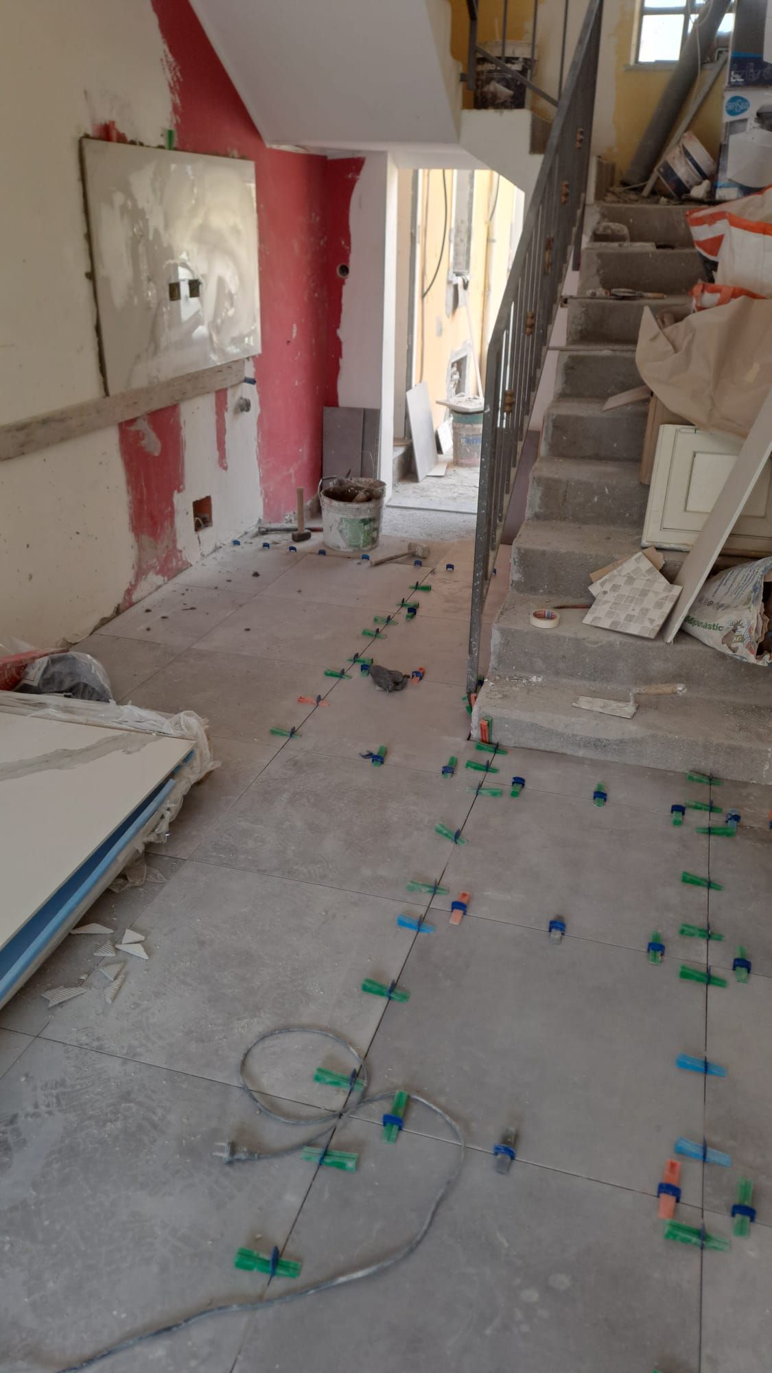 Cantiere interno. Posa delle piastrelle del pavimento, con distanziatori. Sono visibili il muro rosso, le scale e i detriti.