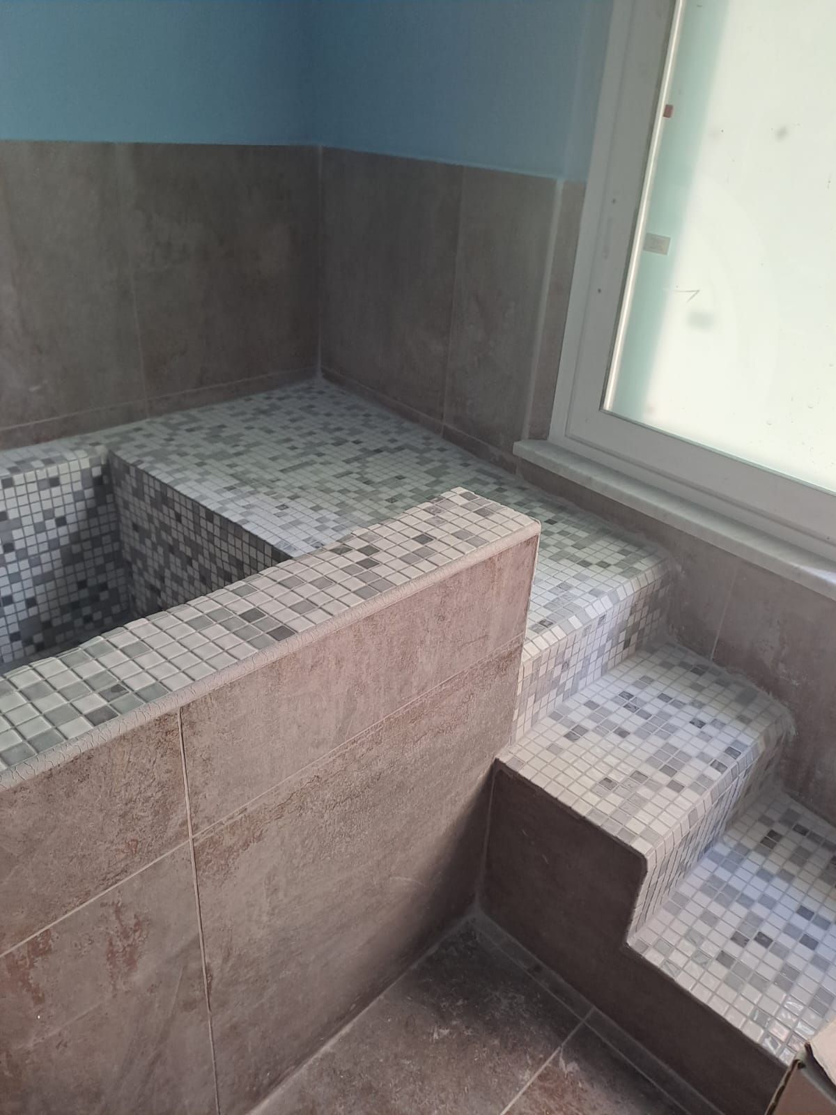 Bagno piastrellato con vasca da bagno integrata e gradini. Pareti marroni con pavimento in piastrelle a mosaico, accanto a una finestra con finiture bianche.