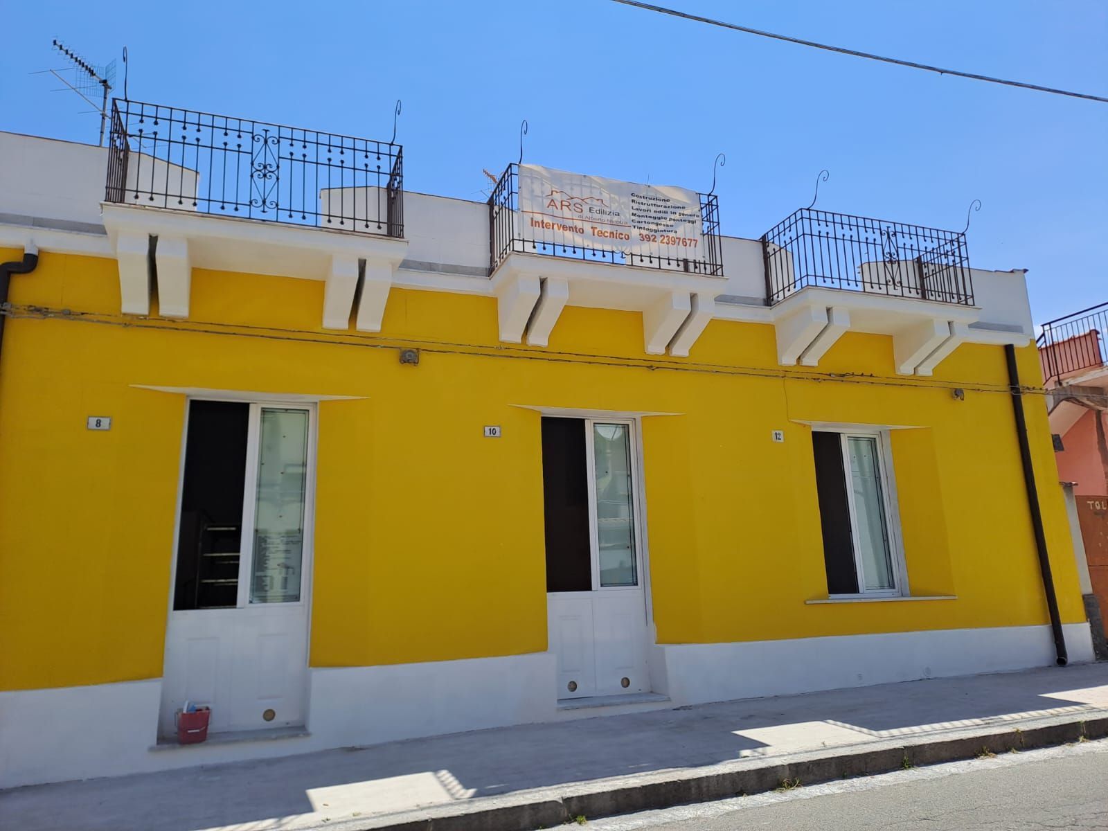 Edificio giallo con finiture e balconi bianchi. Finestre e porte fiancheggiano la facciata.