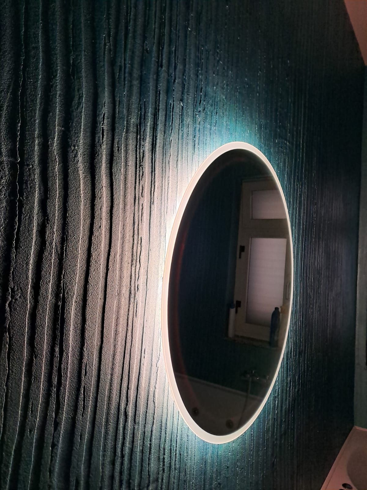 Specchio ovale con bordo illuminato su parete blu testurizzata.