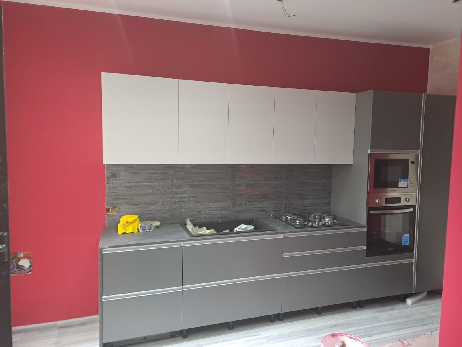 Cucina moderna con mobili grigi, pensili bianchi, parete rossa ed elettrodomestici da incasso.