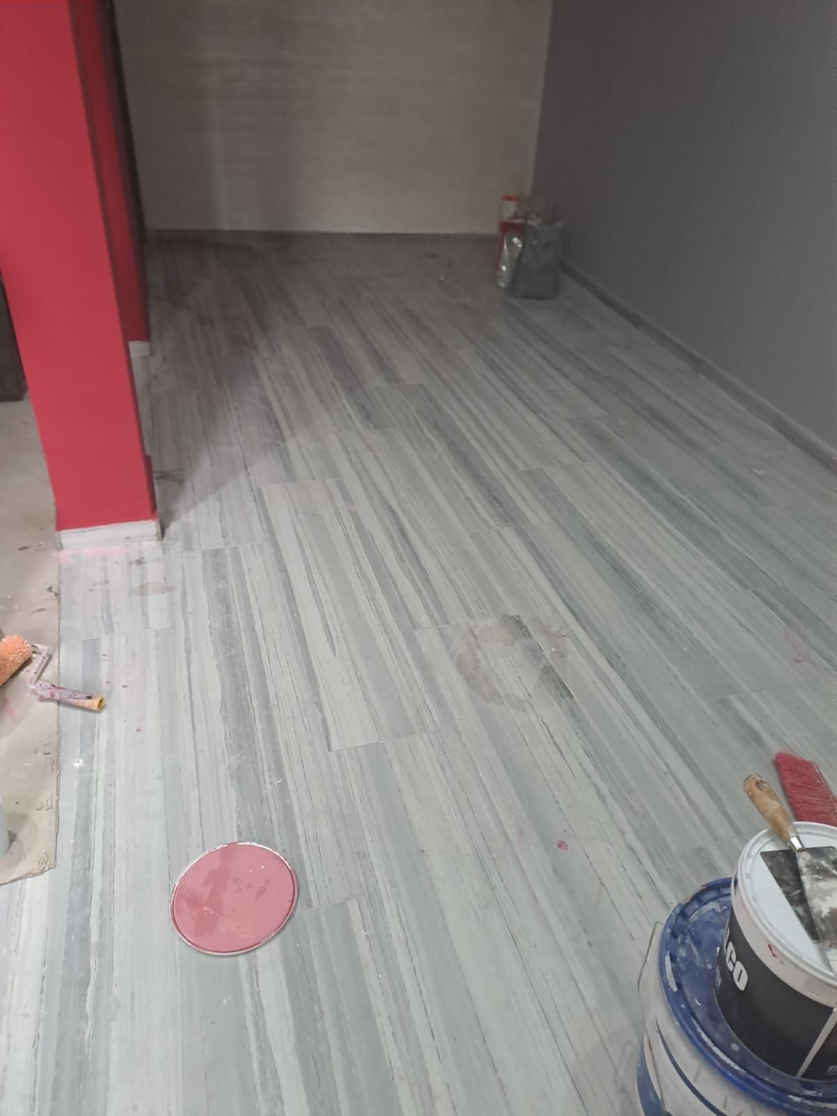 Posa del pavimento: assi grigie e bianche posate in diagonale. Colonna rossa a parete sulla sinistra. Attrezzi da costruzione presenti.