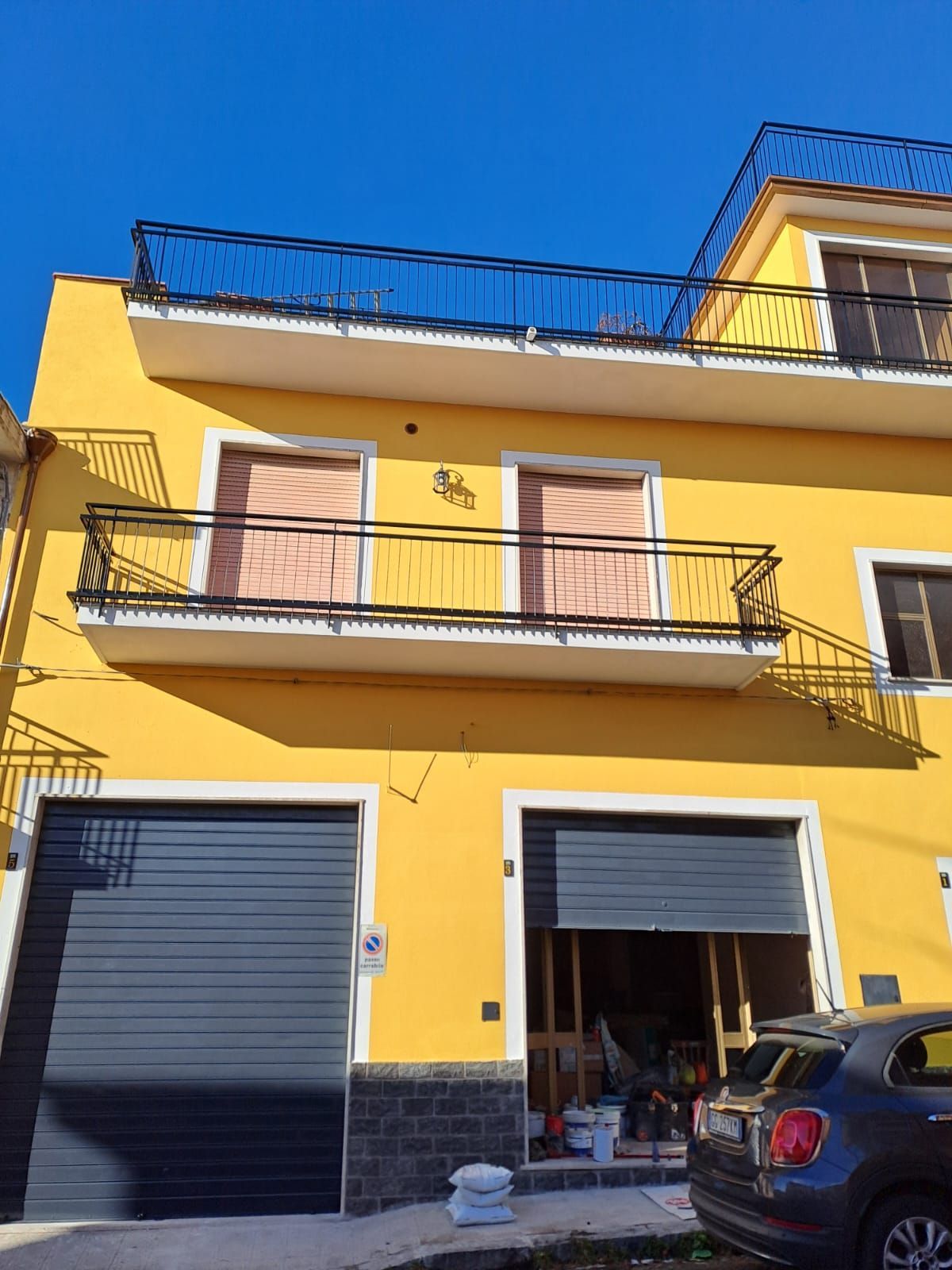 Edificio giallo con porte da garage nere, ringhiere dei balconi e una ringhiera sul tetto sotto un cielo azzurro.