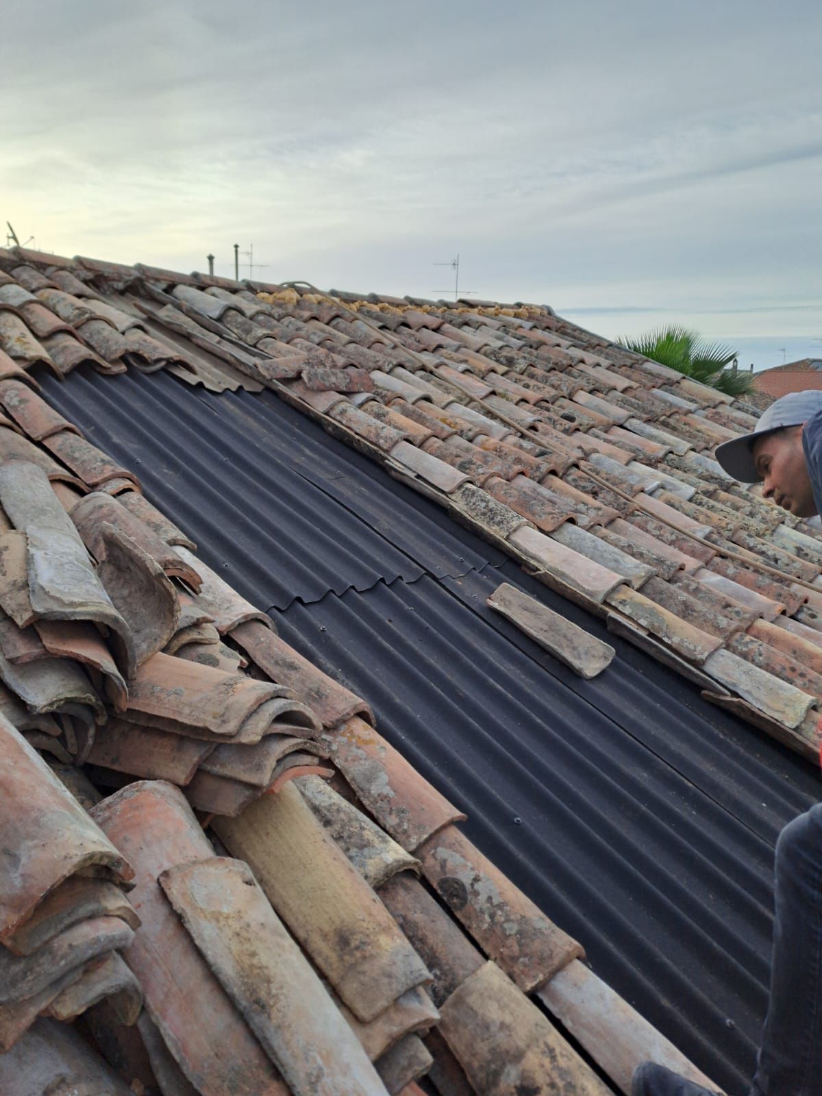 Uomo sul tetto, che sostituisce le tegole di argilla con materiale ondulato nero, cielo coperto.