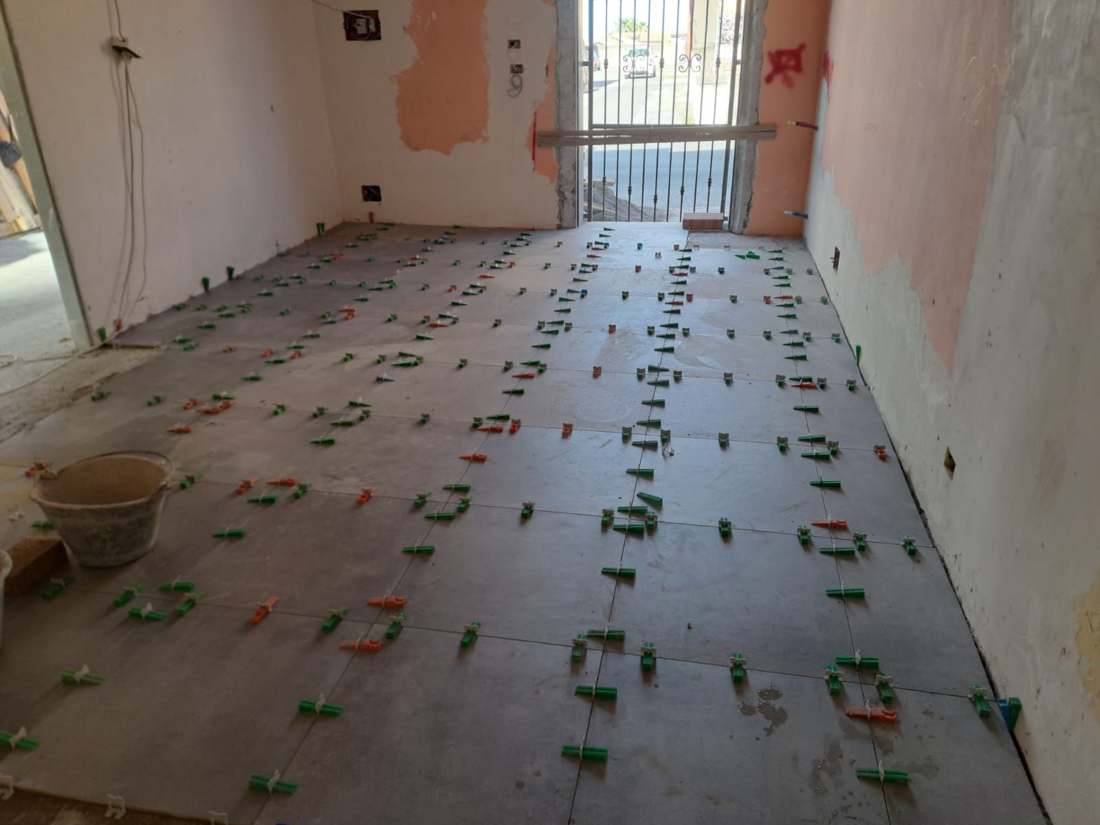 Piastrellatura del pavimento in corso; piastrelle grigie con fermi di livellamento in una stanza con pareti grezze e un cancello di metallo.