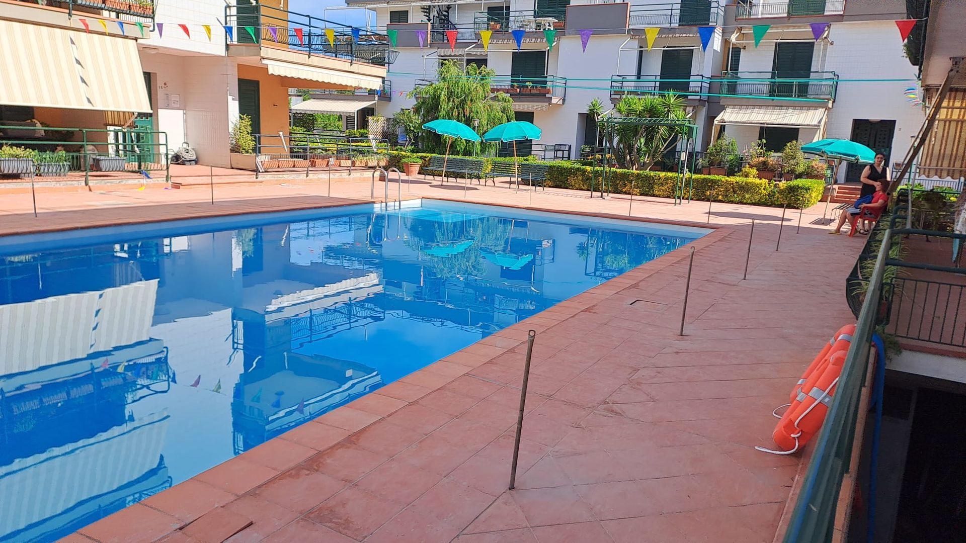 Piscina circondata da edifici. L'acqua azzurra riflette il cielo. Terrazza marrone con salvagente, tavolini e ombrelloni.