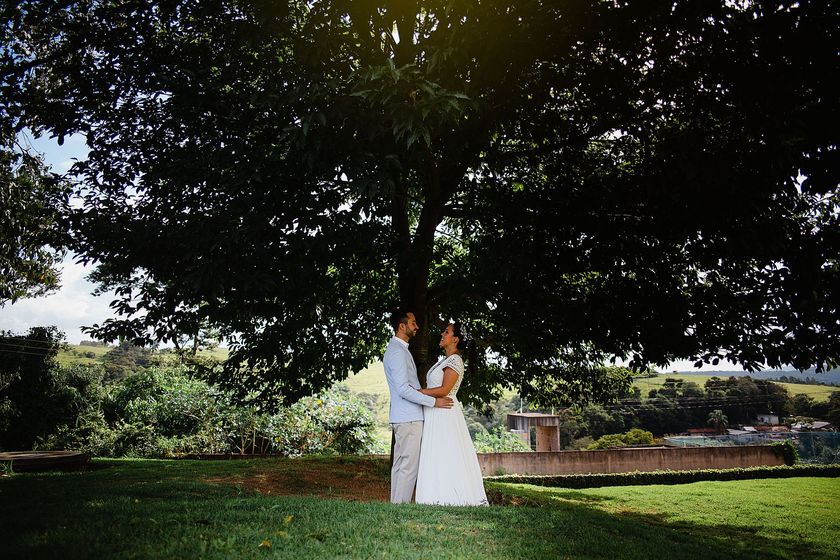 Casal em trajes de casamento brancos, em pé sob uma grande árvore em um gramado ao ar livre.