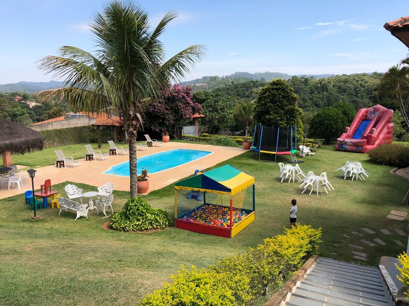Quintal com piscina, palmeira, mesas de piquenique, toldo colorido e escorregador rosa em um dia ensolarado.
