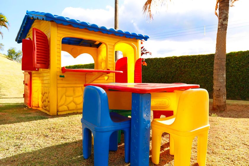 Casinha de plástico colorida, mesa e cadeiras infantis na grama de um quintal ensolarado.