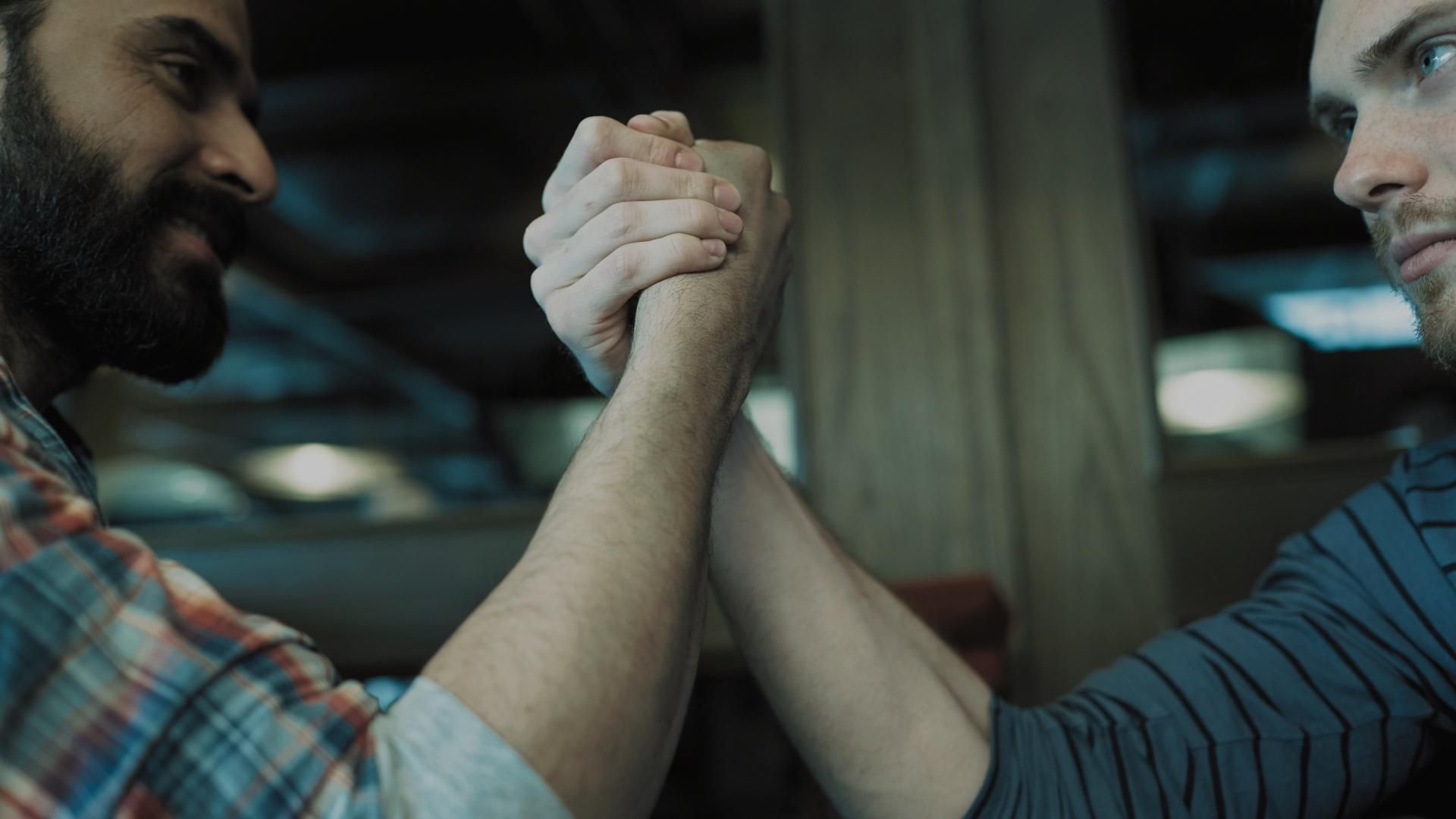 Two men arm wrestling, indoors. One with a beard, plaid shirt; the other with a striped shirt.