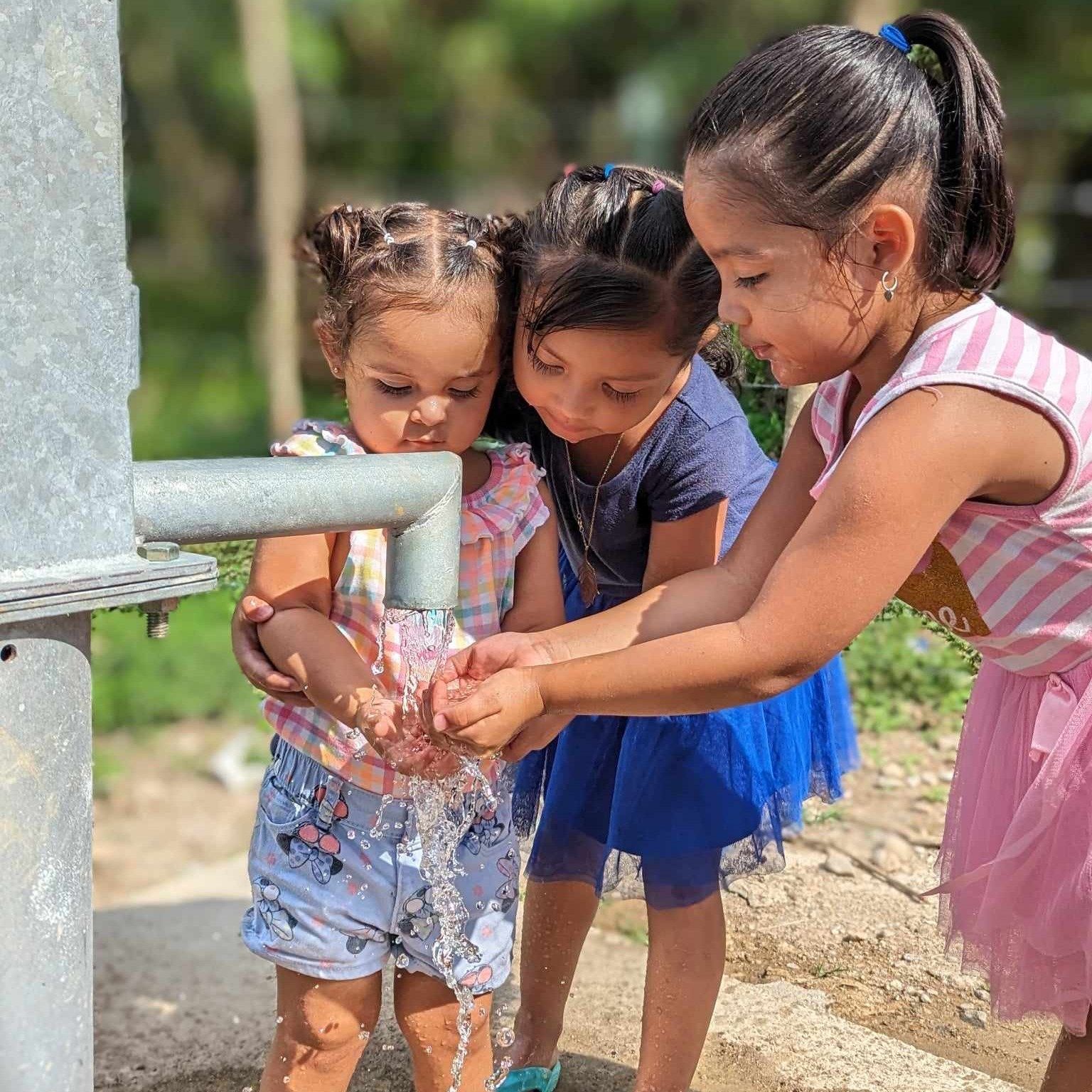 Three little girls are washing their hands under a water pump.