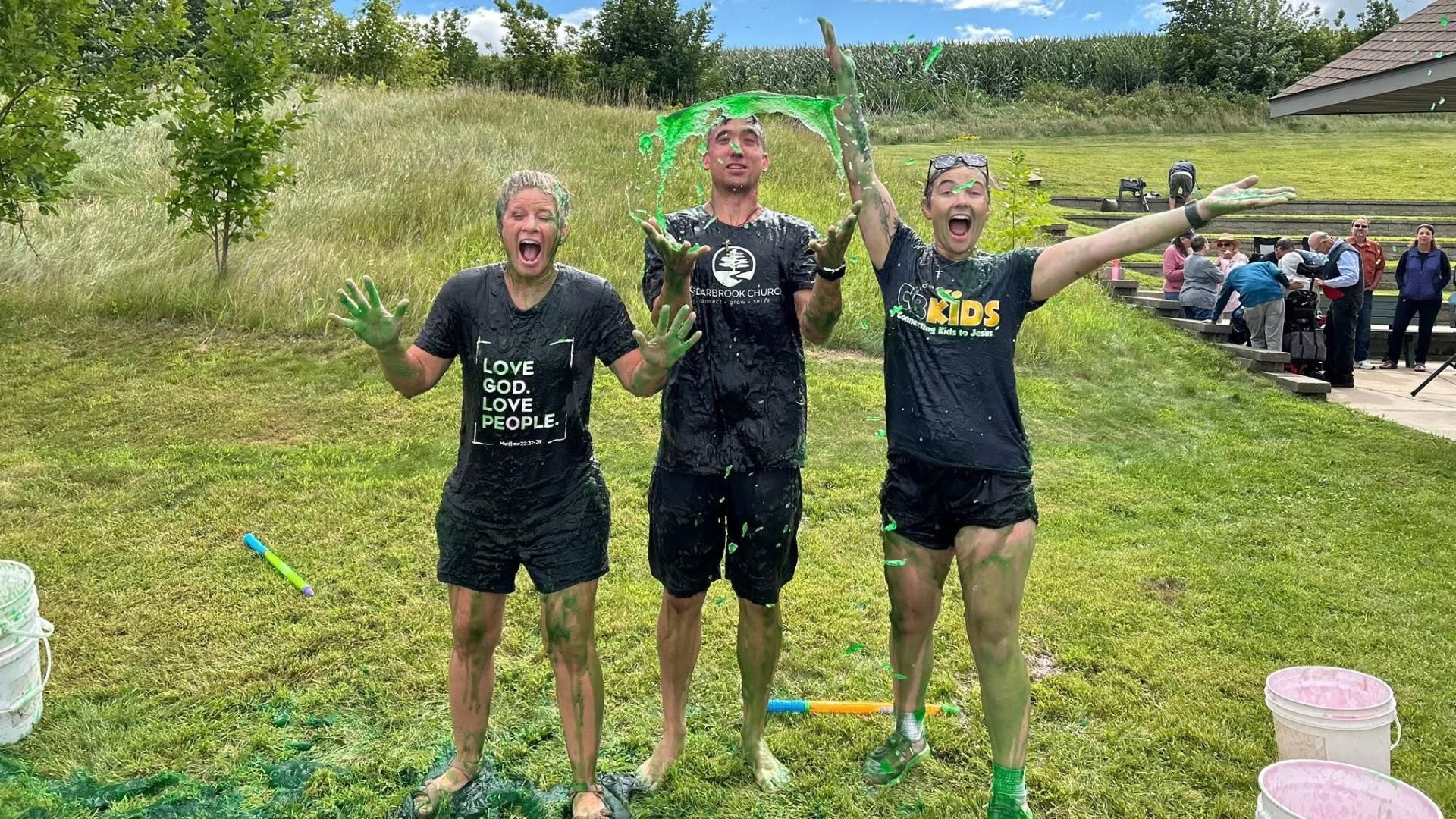Three people covered in green slime outdoors, smiling and celebrating.
