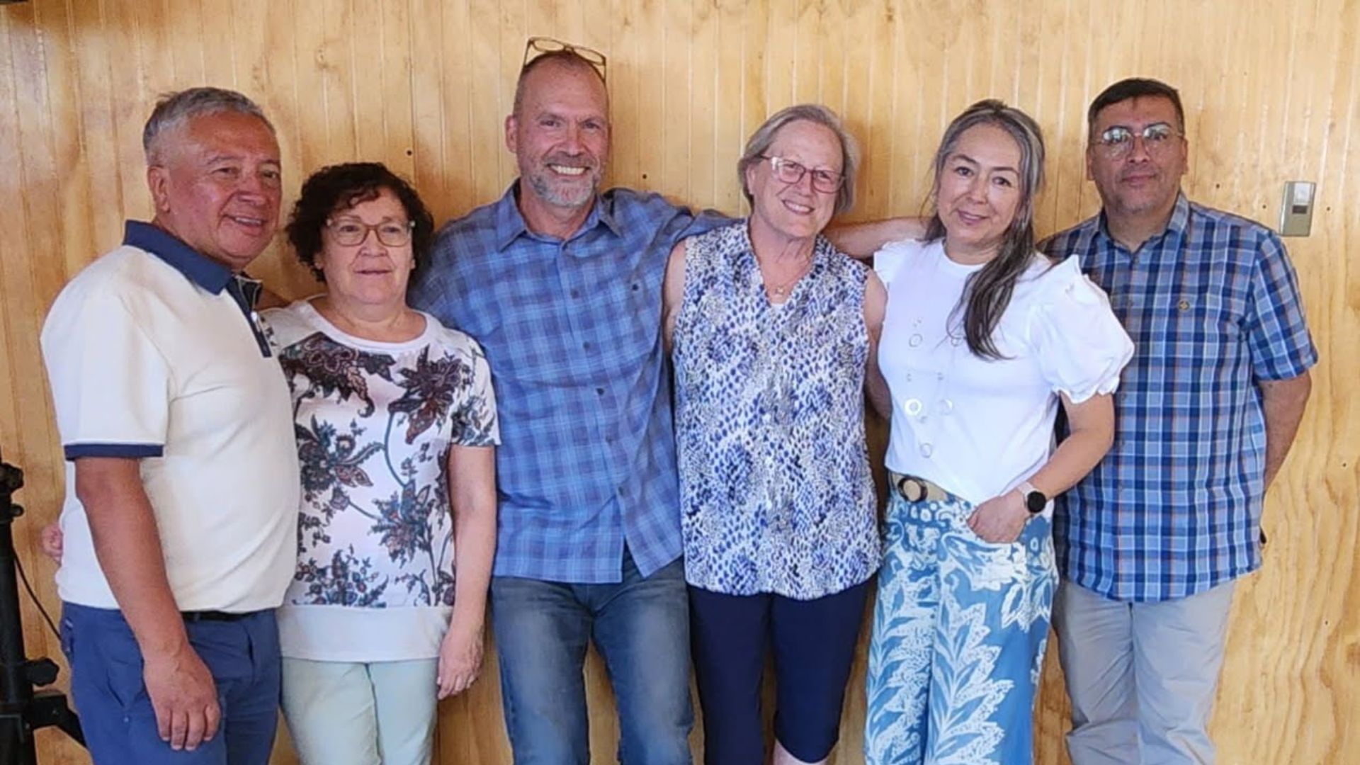 Six people standing together, arms around each other, in front of wood paneling.
