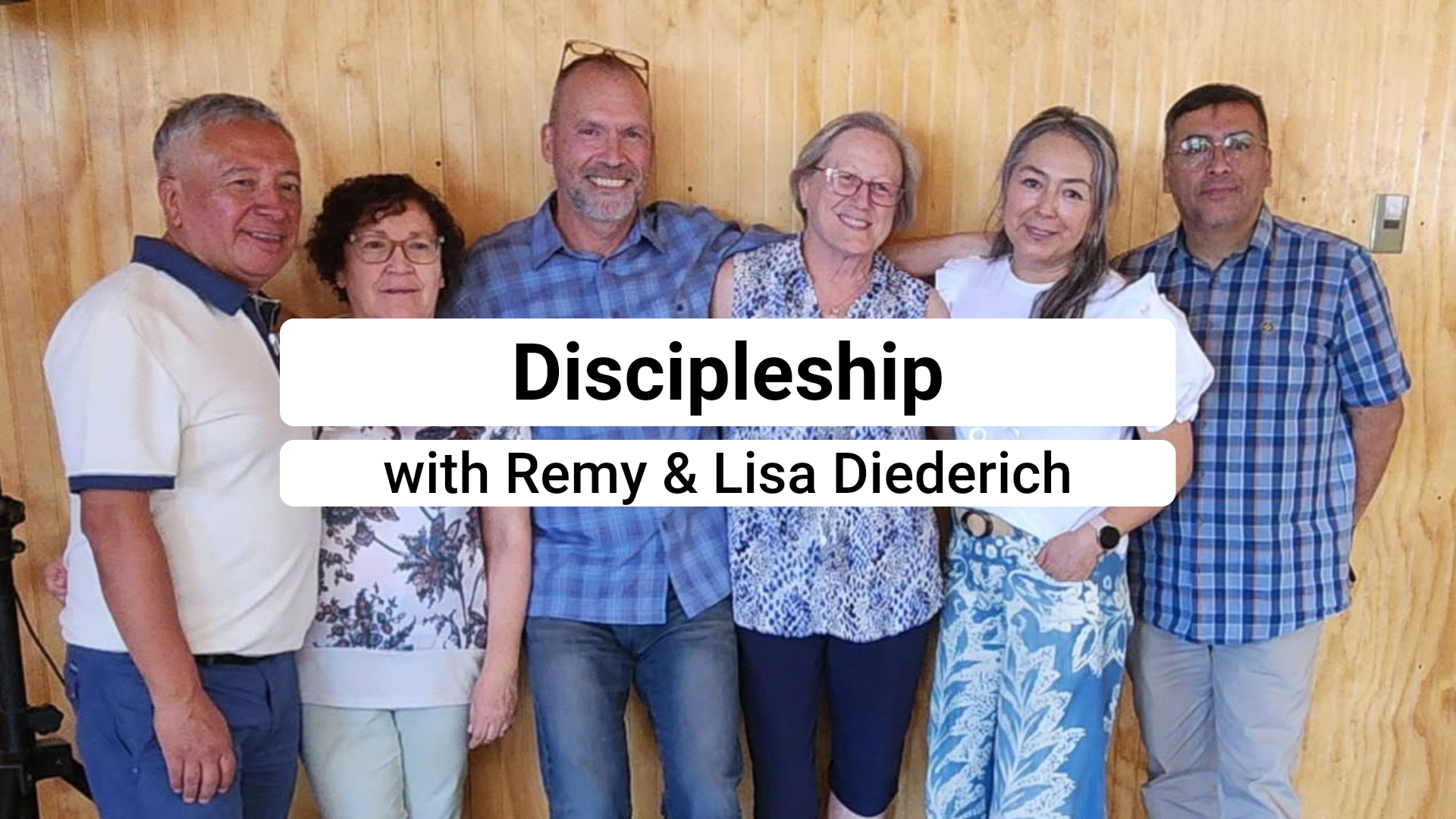 Six people standing in a row against a wood-paneled wall. A banner in the center reads 