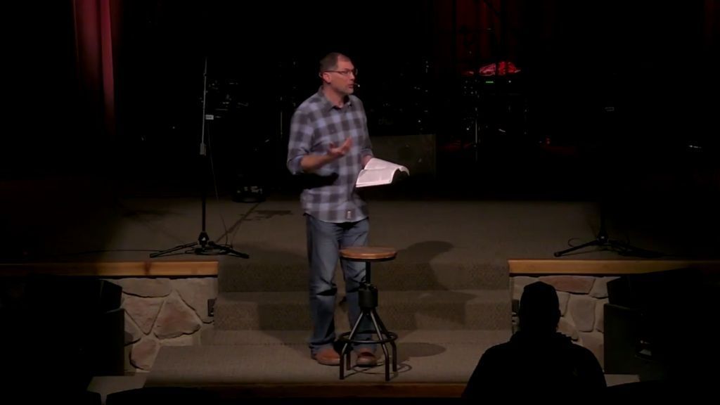 Michael Behm speaking on a stage, holding a Bible, wearing glasses, and a plaid shirt. Dark background.