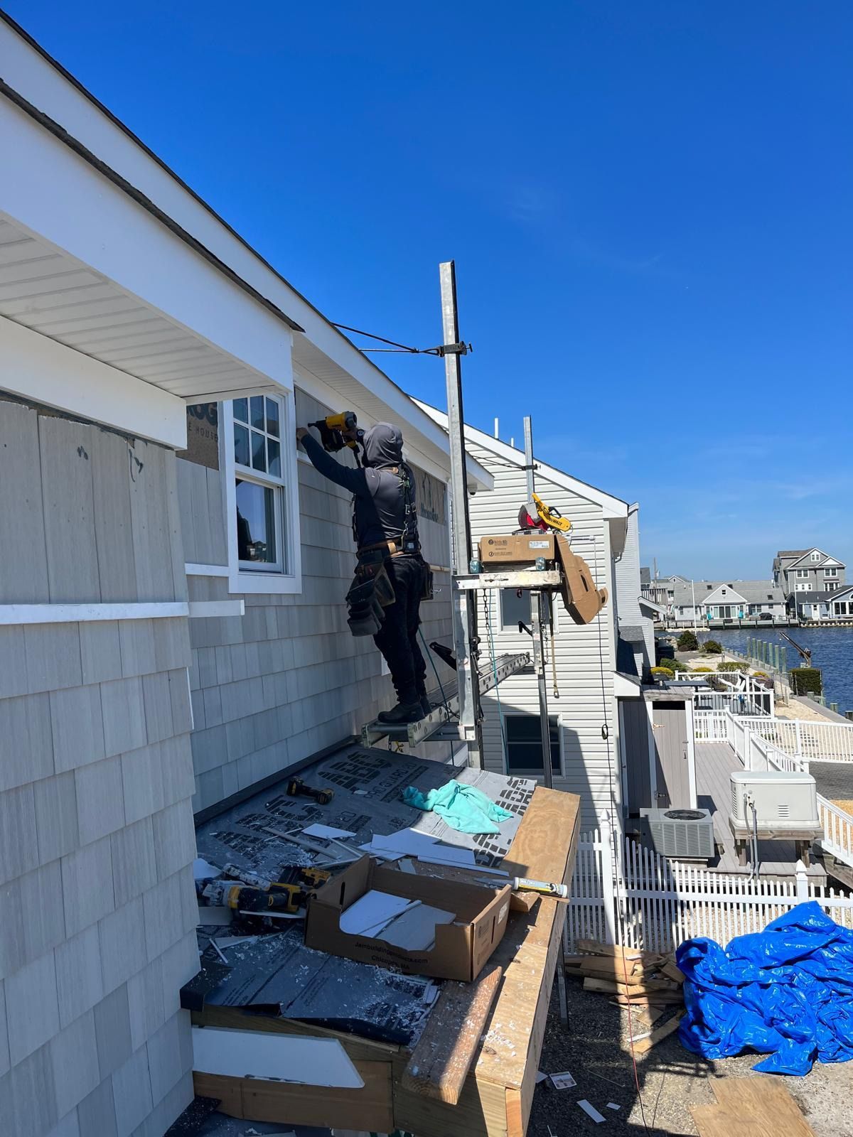 Person on scaffolding installing siding on a house, sunny day, water in the background.
